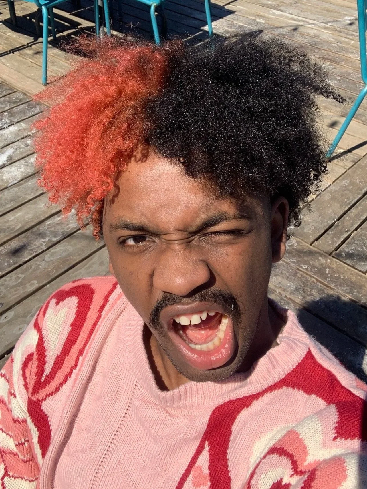 A  beautiful young man with curly half black, half pink hair taking a selfie outdoors on a sunny day, wearing a pink and red patterned sweater, with a playful expression and winking eye.
