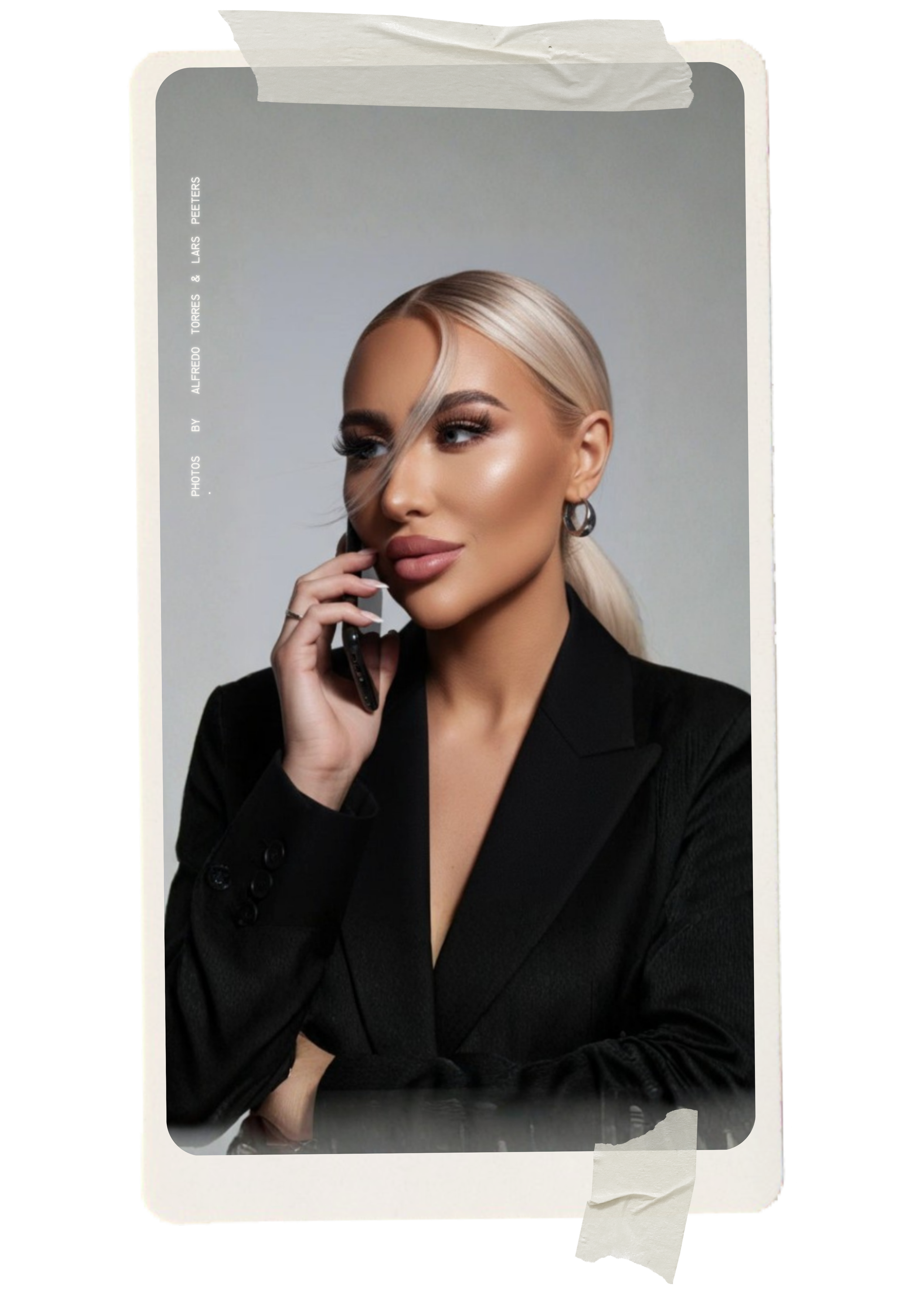 A woman with platinum blonde hair styled in a sleek ponytail, wearing a black blazer, holding a phone to her ear, and posing against a gray background.