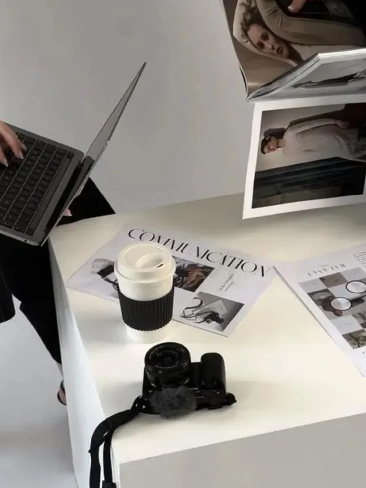 A white table with a laptop, a camera, a disposable coffee cup, and magazine pages on it. There are photos of a woman in various poses, some of which are partially visible. A person is standing nearby.