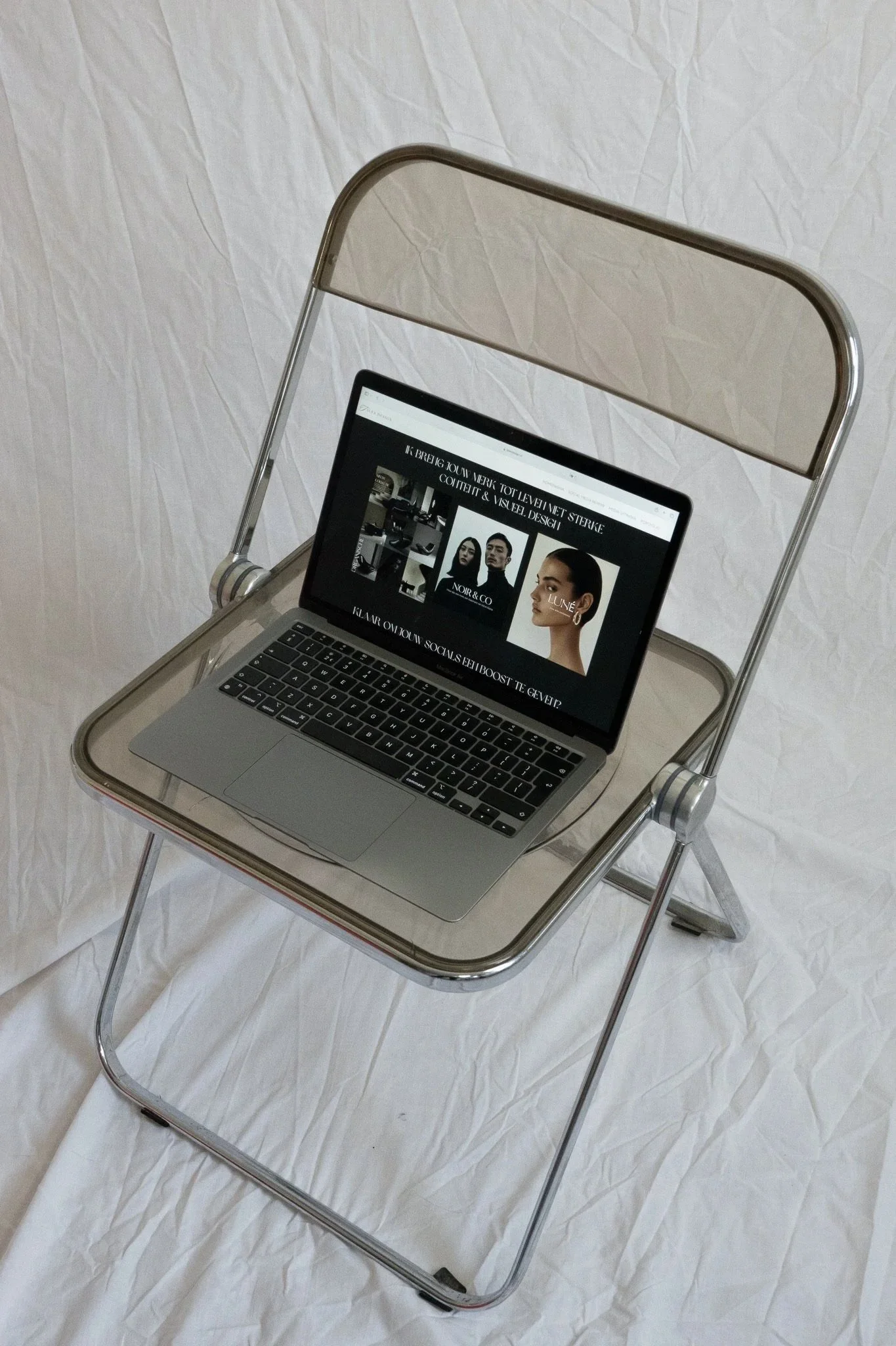 A silver folding cart with a laptop on top, displaying a fashion website with images of models and jewelry. The cart is set against a wrinkled white background.