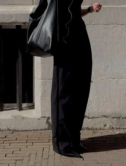 A person standing outdoors, wearing black pants, a black top, and holding a black bag, with crutches in hand, against a concrete wall.