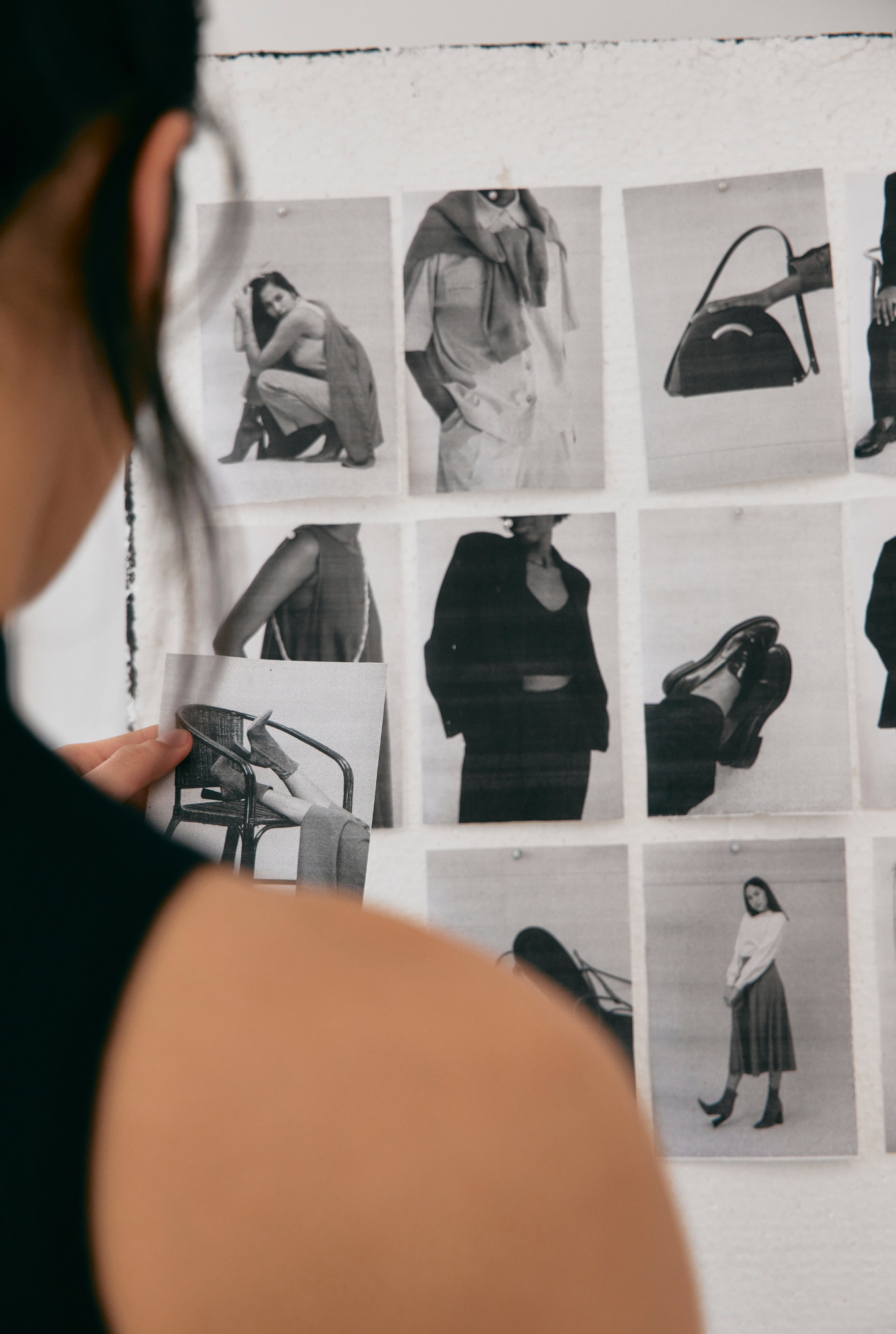 A person holding a small photograph in front of a wall with black-and-white fashion magazine photos, including women posing with handbags and shoes.