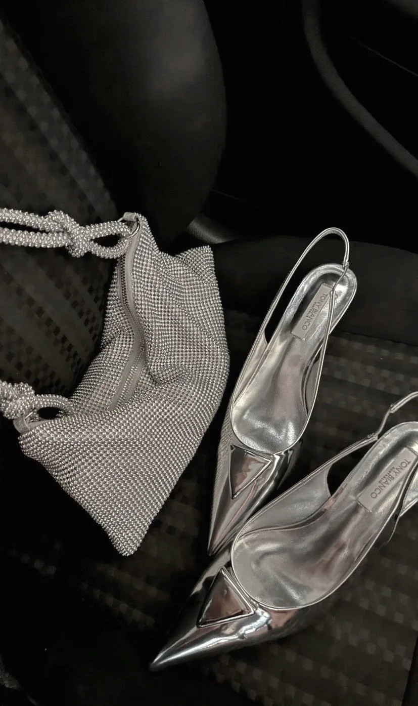 A pair of shiny silver high-heeled shoes and a matching silver sparkling bag with a zipper and a knotted strap are placed on a black car seat.