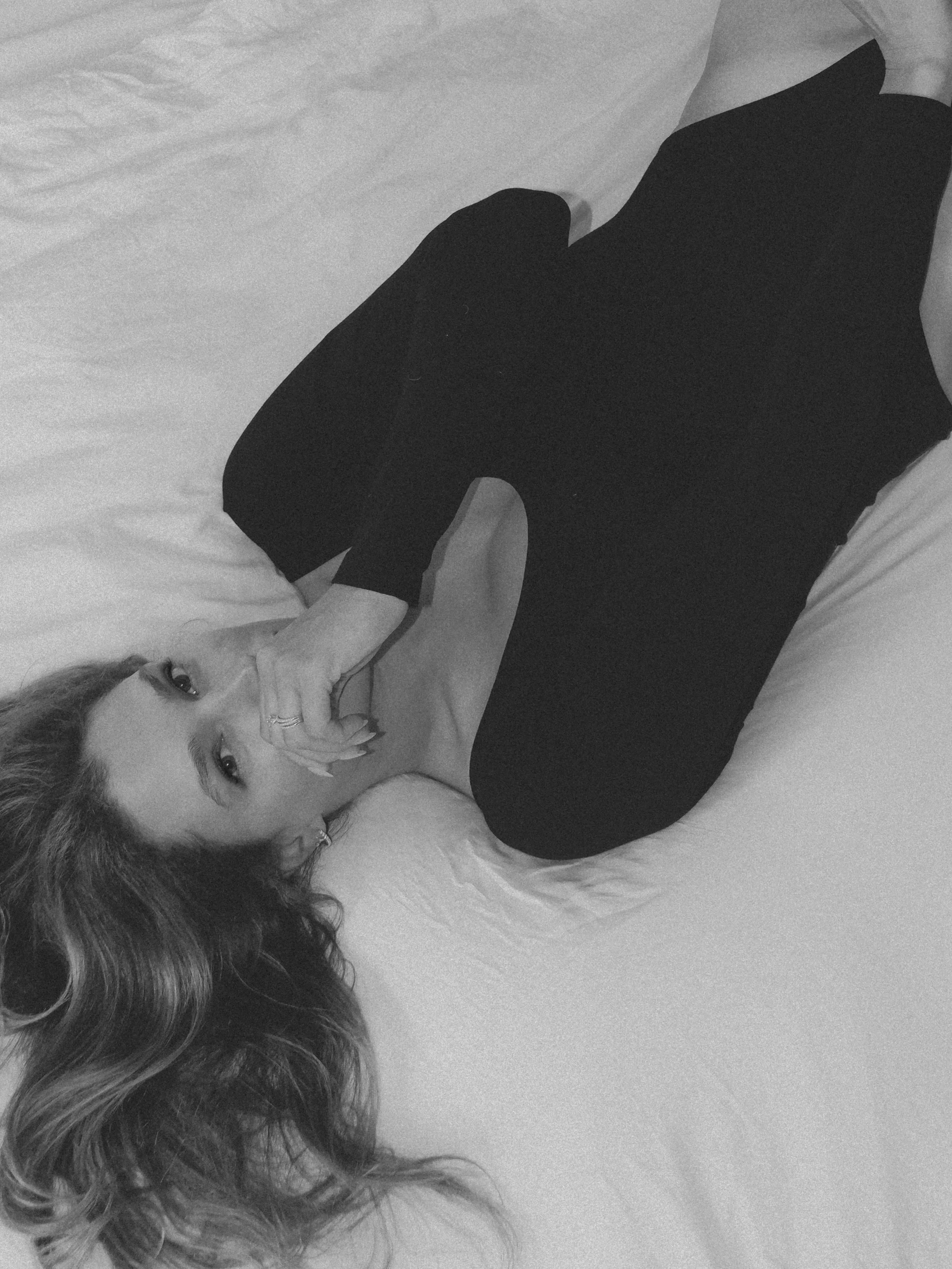 A woman lying on a bed with long curly hair, hand near her face, looking at the camera in black and white.