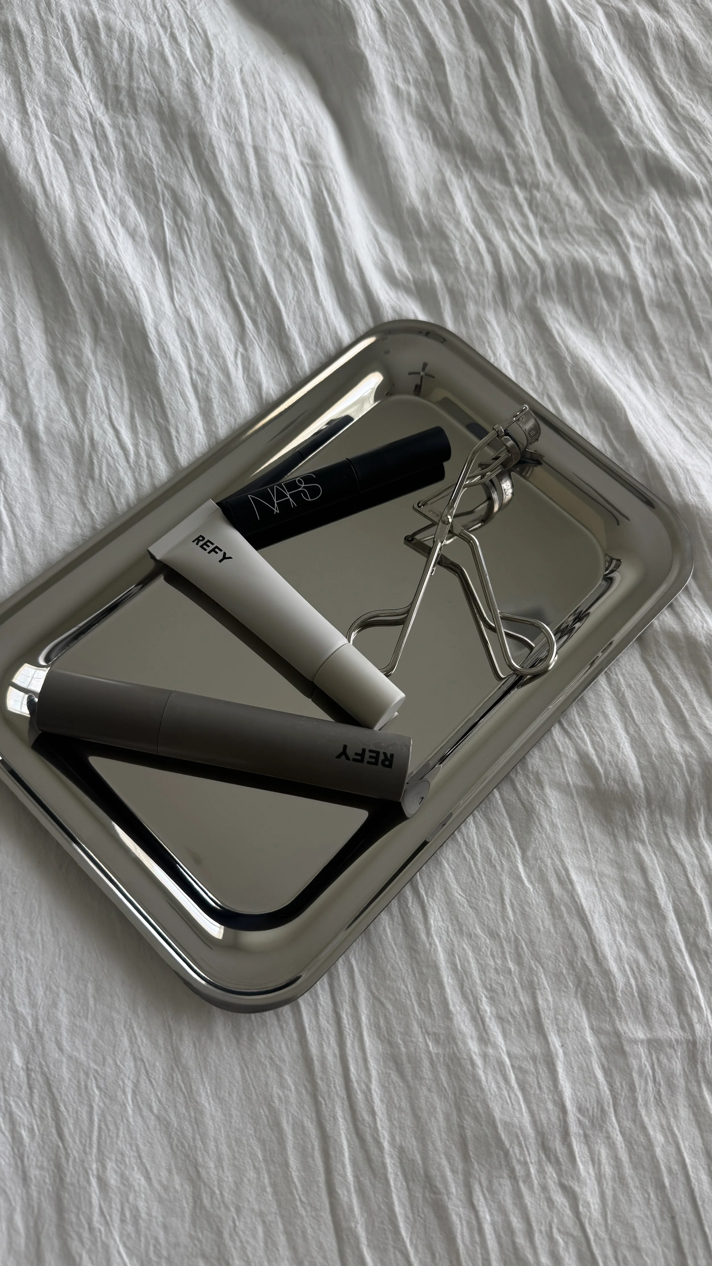 A silver tray with a black tube labeled 'NARS,' a white tube labeled 'REY,' a gray tube, and an eyelash curler resting on a white textured fabric surface.