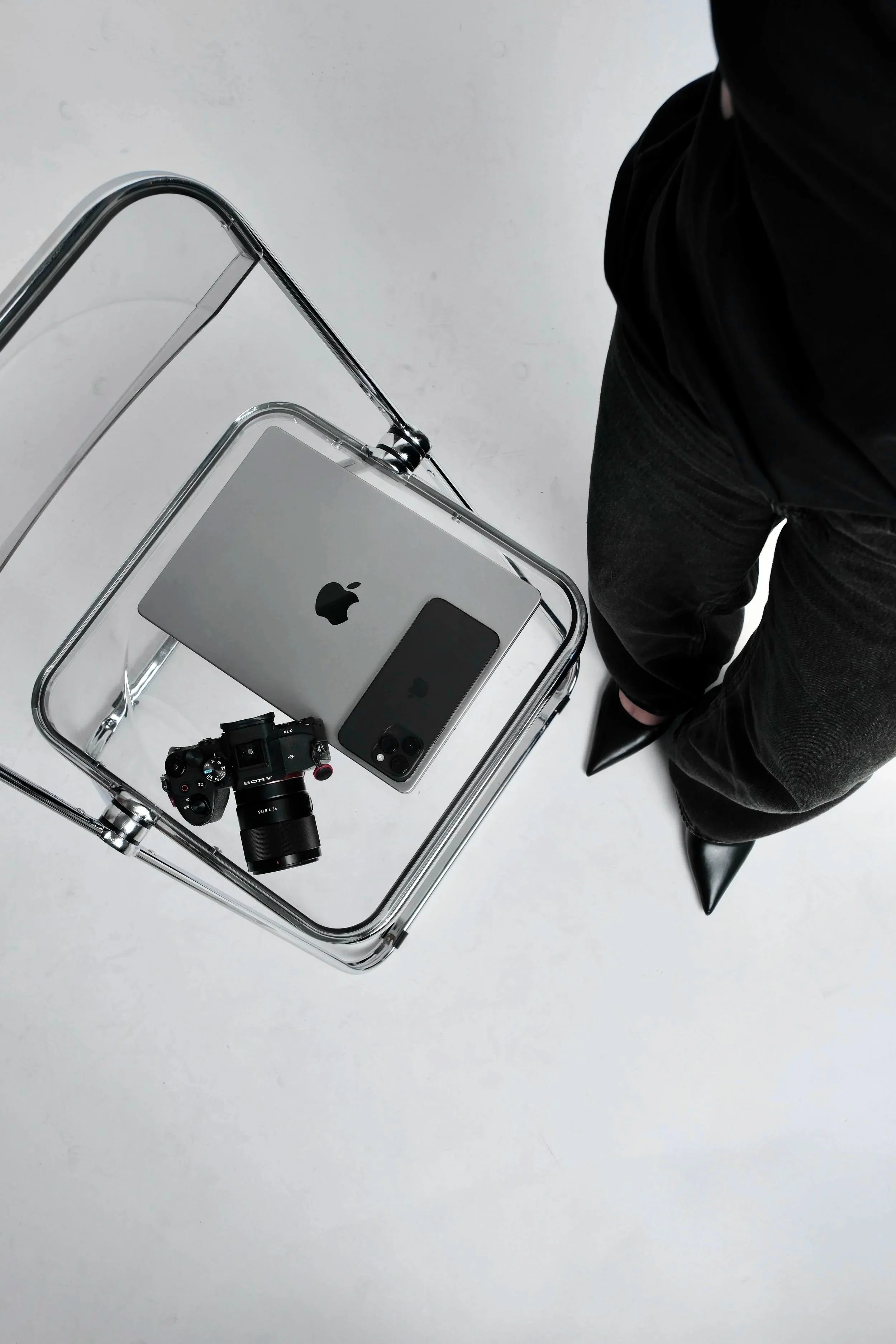 Top-down view of a glass table with Apple products, including an iPhone, a MacBook, and an iPad, along with a Sony camera, on a white floor, with a person dressed in black standing next to it.