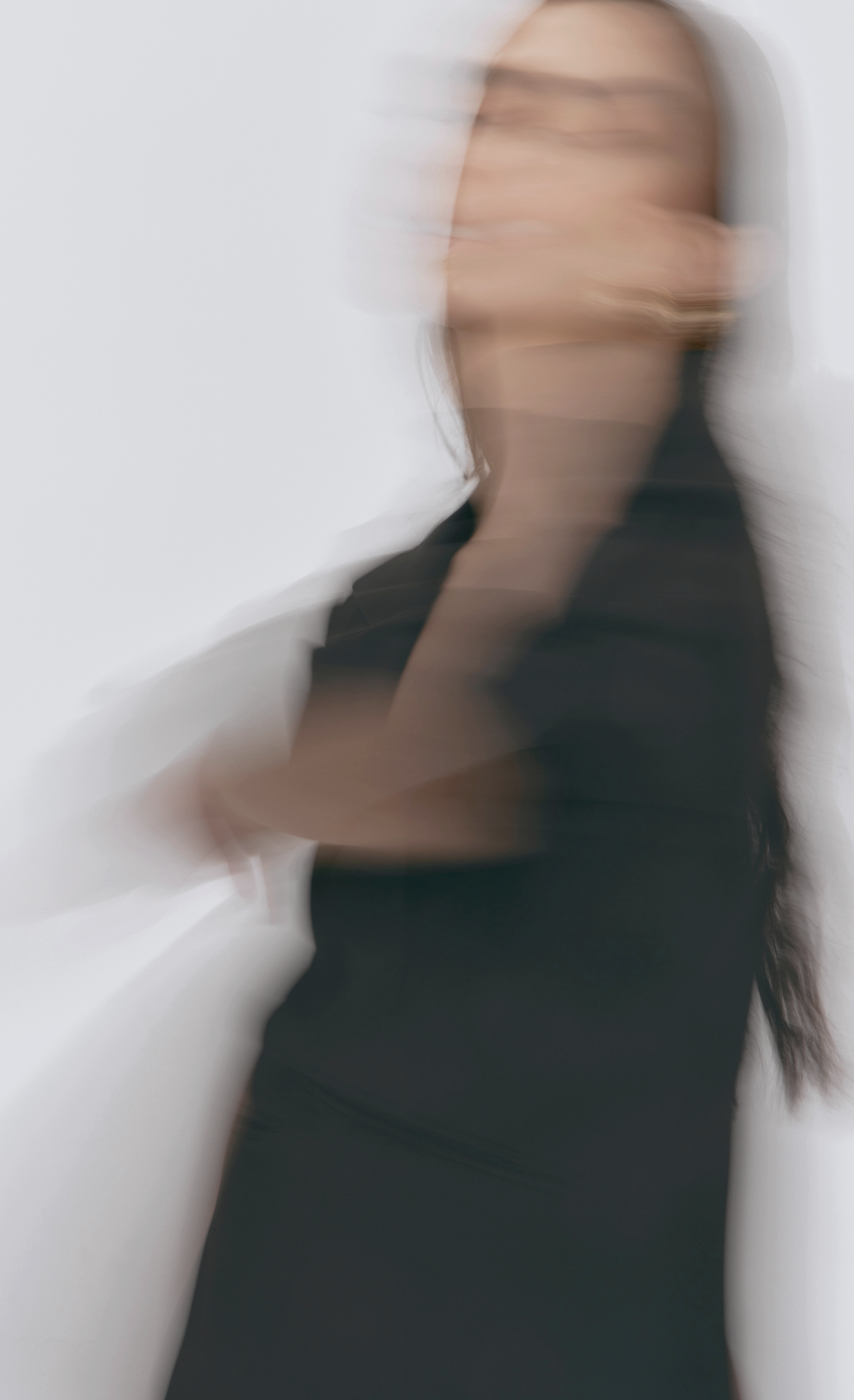 Blurred image of a woman with long dark hair, dressed in black, standing against a white background.