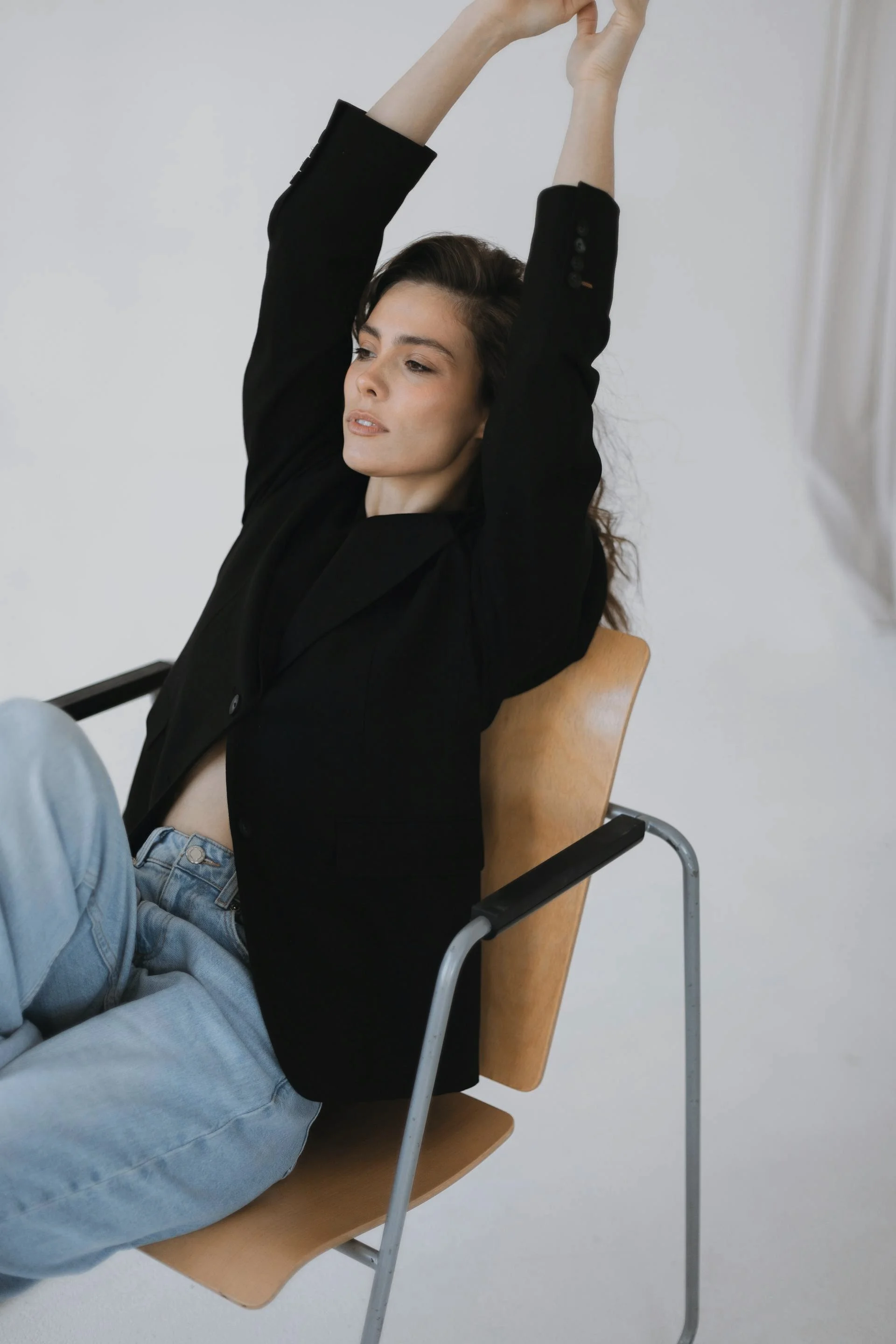 A woman with dark brown hair wearing a black jacket and gray jeans, sitting in a chair with her arms raised above her head, stretching in a plain room with a white wall and a beige curtain.