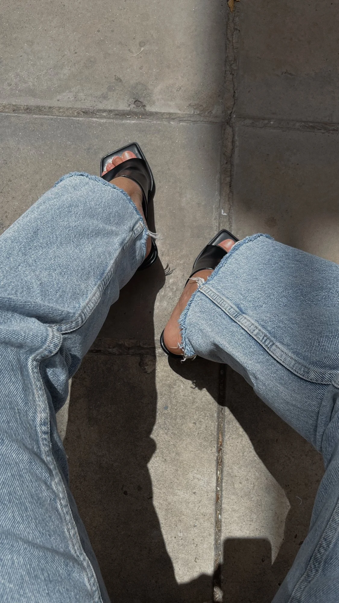 View of a person's legs wearing ripped blue jeans and black flip-flops, sitting on a concrete sidewalk with shadows cast on the ground.