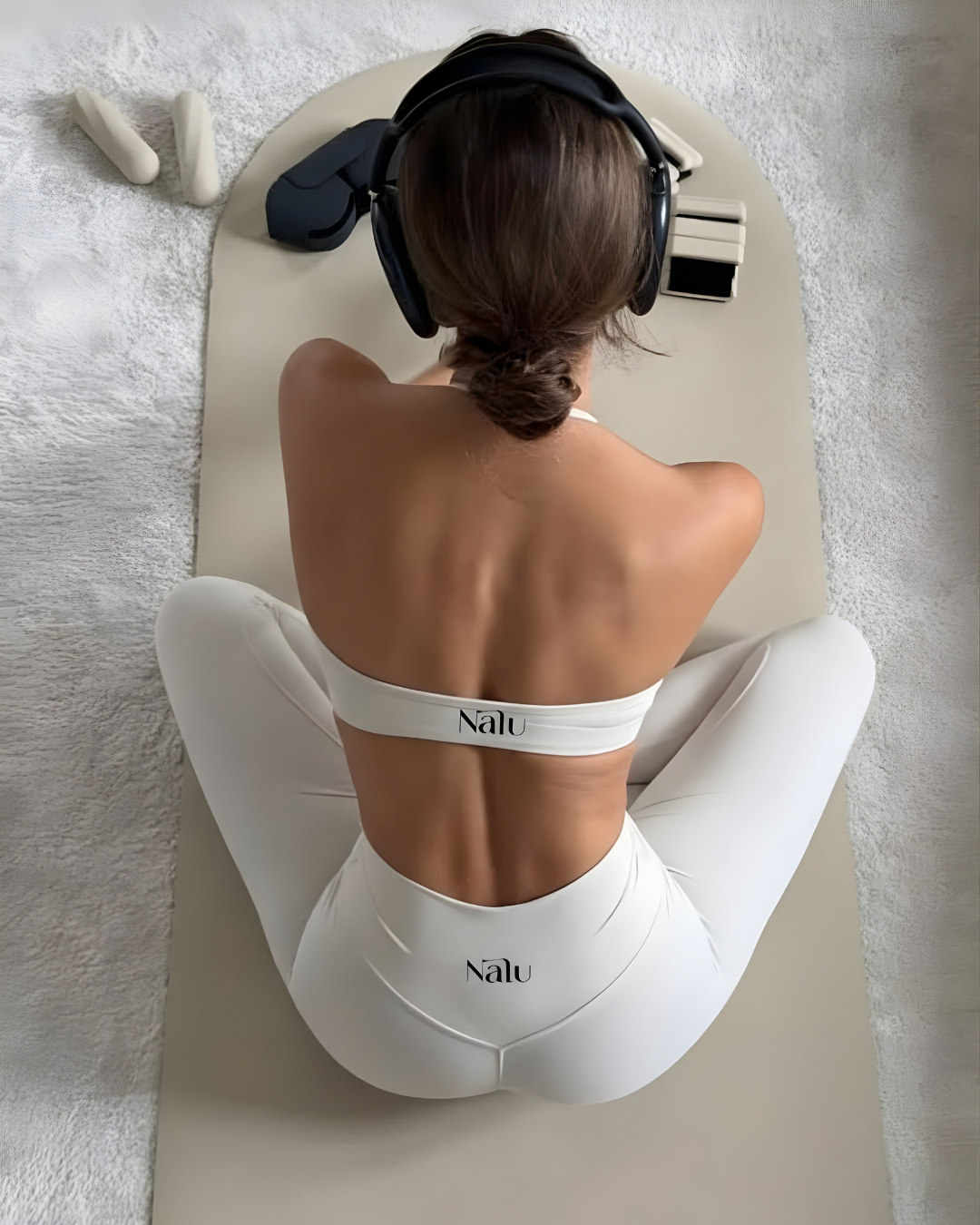 A woman with brown hair tied back, wearing white athletic wear with the word 'Nalu' on the back, is sitting cross-legged on a mat. She is wearing large black headphones and appears to be engaged in a meditation or relaxation activity. There are some white objects near her on the mat, and the background is a light-colored carpet.