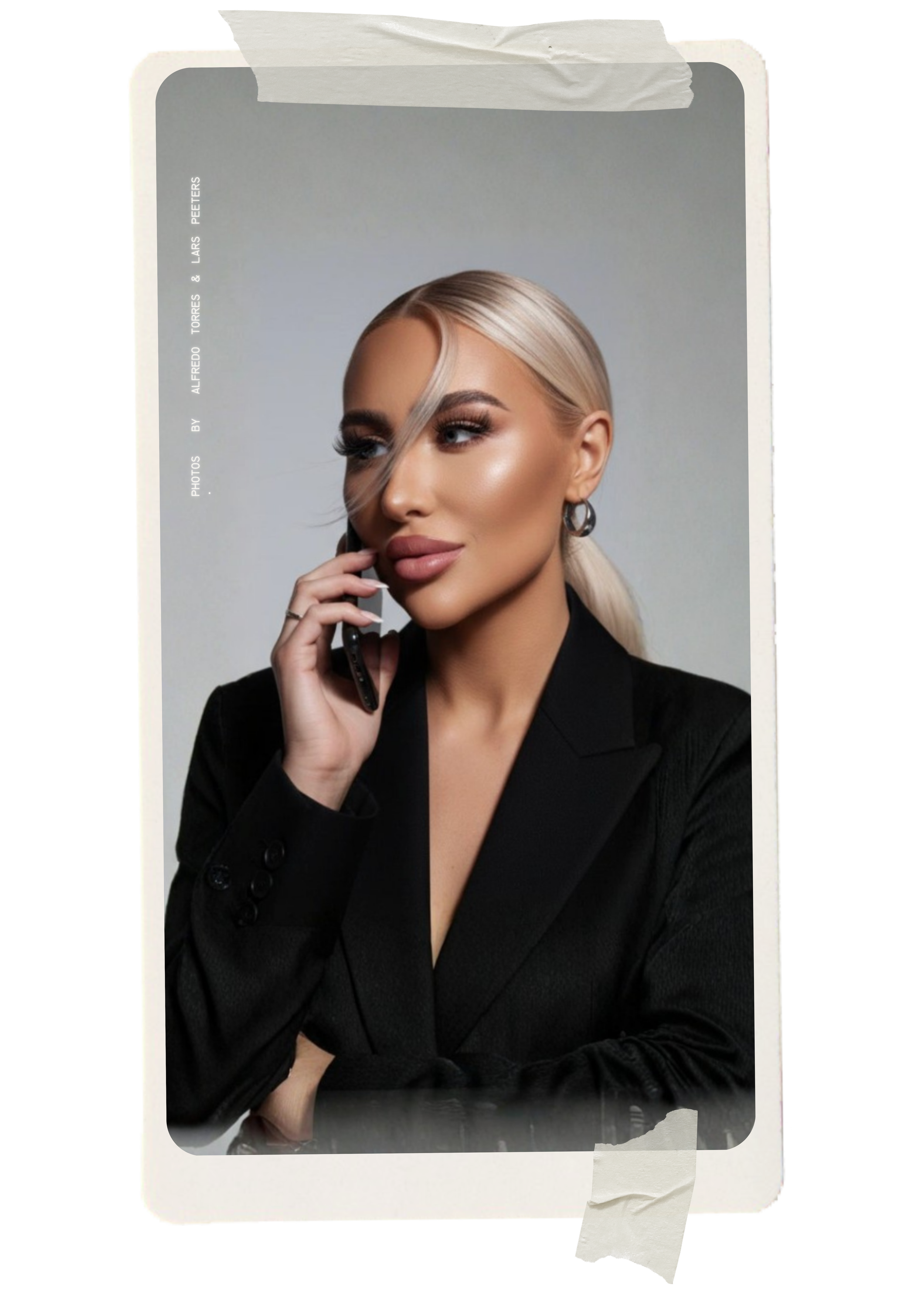 A woman with blonde hair styled back, wearing a black blazer, holding a phone to her ear, with makeup and hoop earrings, against a plain background.