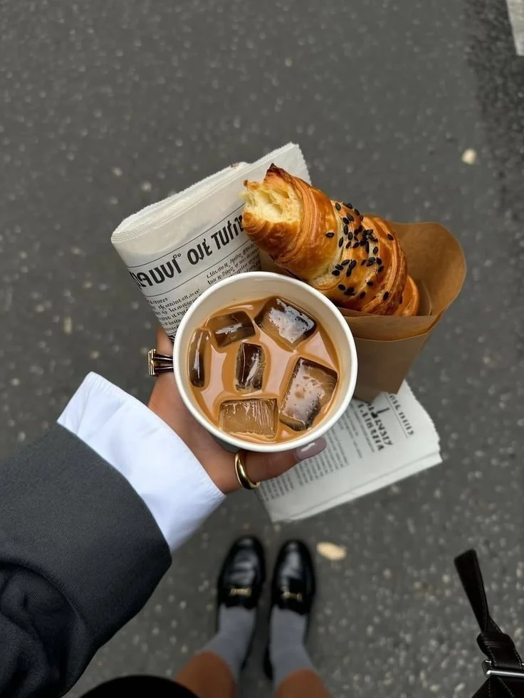 A person holding a cup of iced coffee, a croissant with sesame seeds in a paper bag, and wrapped newspaper or paper packaging, standing on a gray asphalt street.