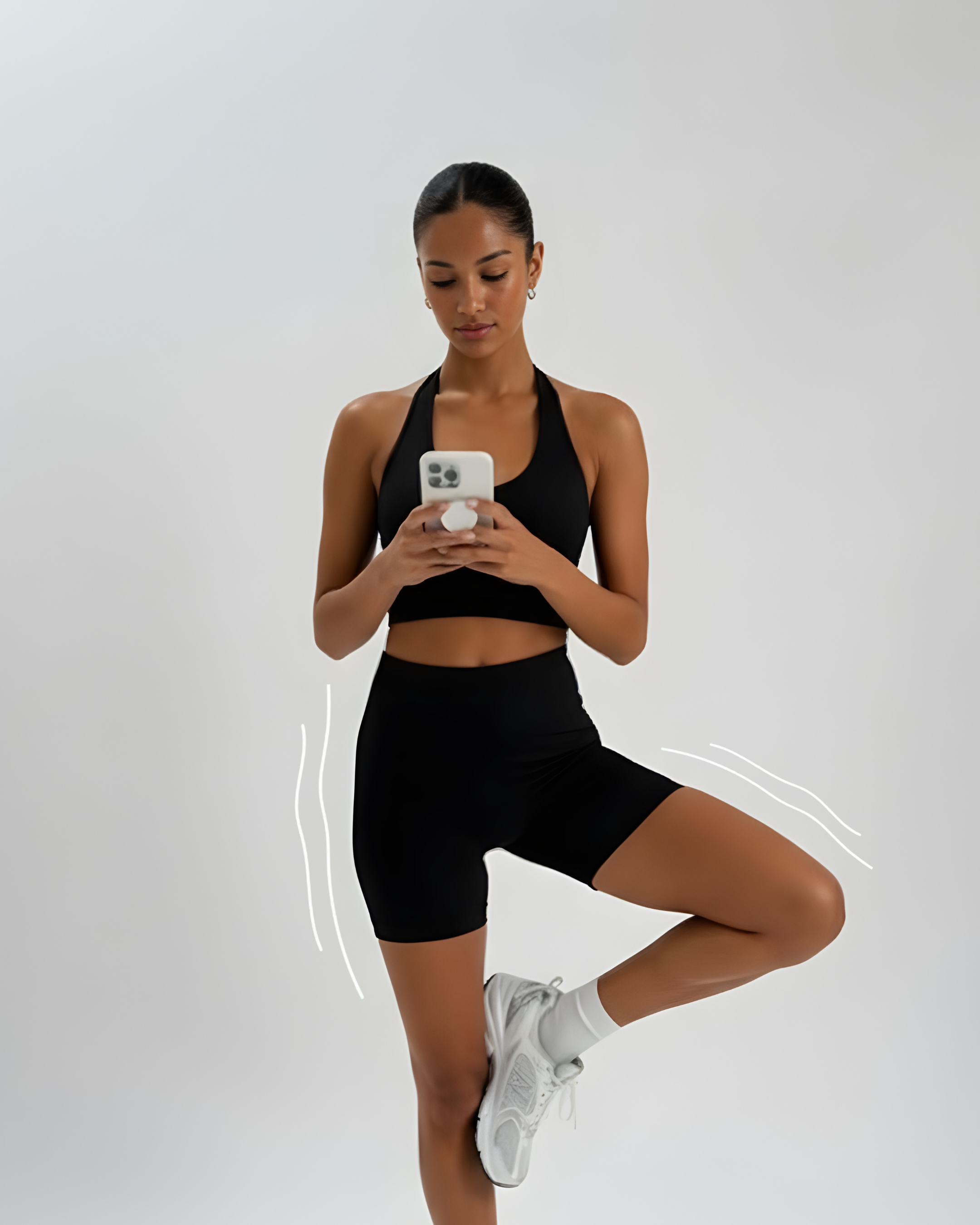 A young woman in athletic wear, including a black sports bra and shorts, white sneakers, and earrings, is performing a yoga pose against a plain white background. She is standing on one leg, with her other knee lifted and her foot resting on her thigh, while she looks at her smartphone.