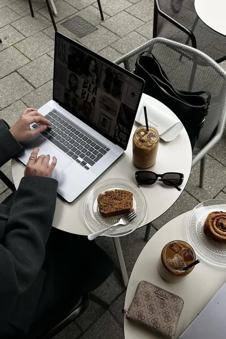 A person working on a laptop at an outdoor café table with iced coffee, sunglasses, a slice of cake, and a second iced coffee, with another table nearby.