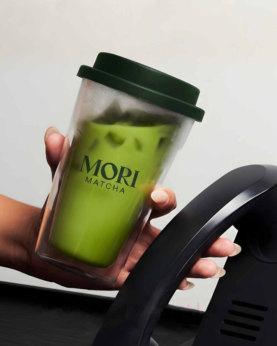 A person's hand is holding a clear to-go cup filled with green matcha latte, with a green lid, in front of a black telephone on a desk, with a plain white wall in the background.
