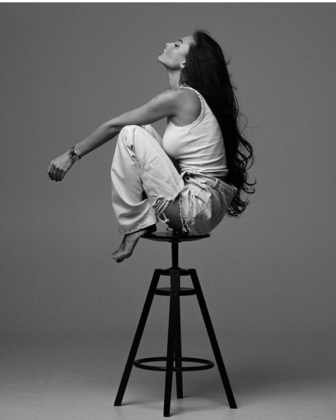Black and white photo of a woman sitting on a high stool against a plain background, with her head tilted back and eyes closed, wearing a sleeveless top and ripped jeans with long, flowing hair.