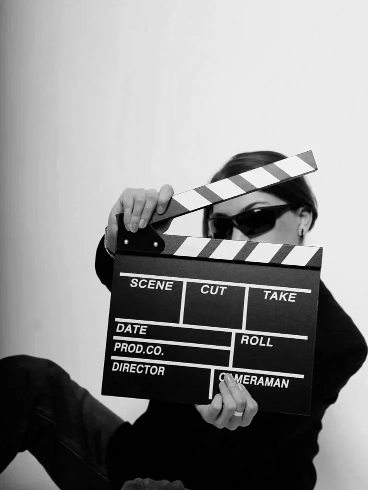A person with dark hair, wearing sunglasses and dark clothing, holding a black and white film slate in front of their face, partially obscuring it. The person is sitting against a plain wall.