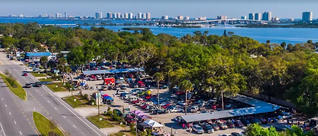 A large parking lot filled with cars and tents, with trees around it, near a road; a City skyline and water in the background.