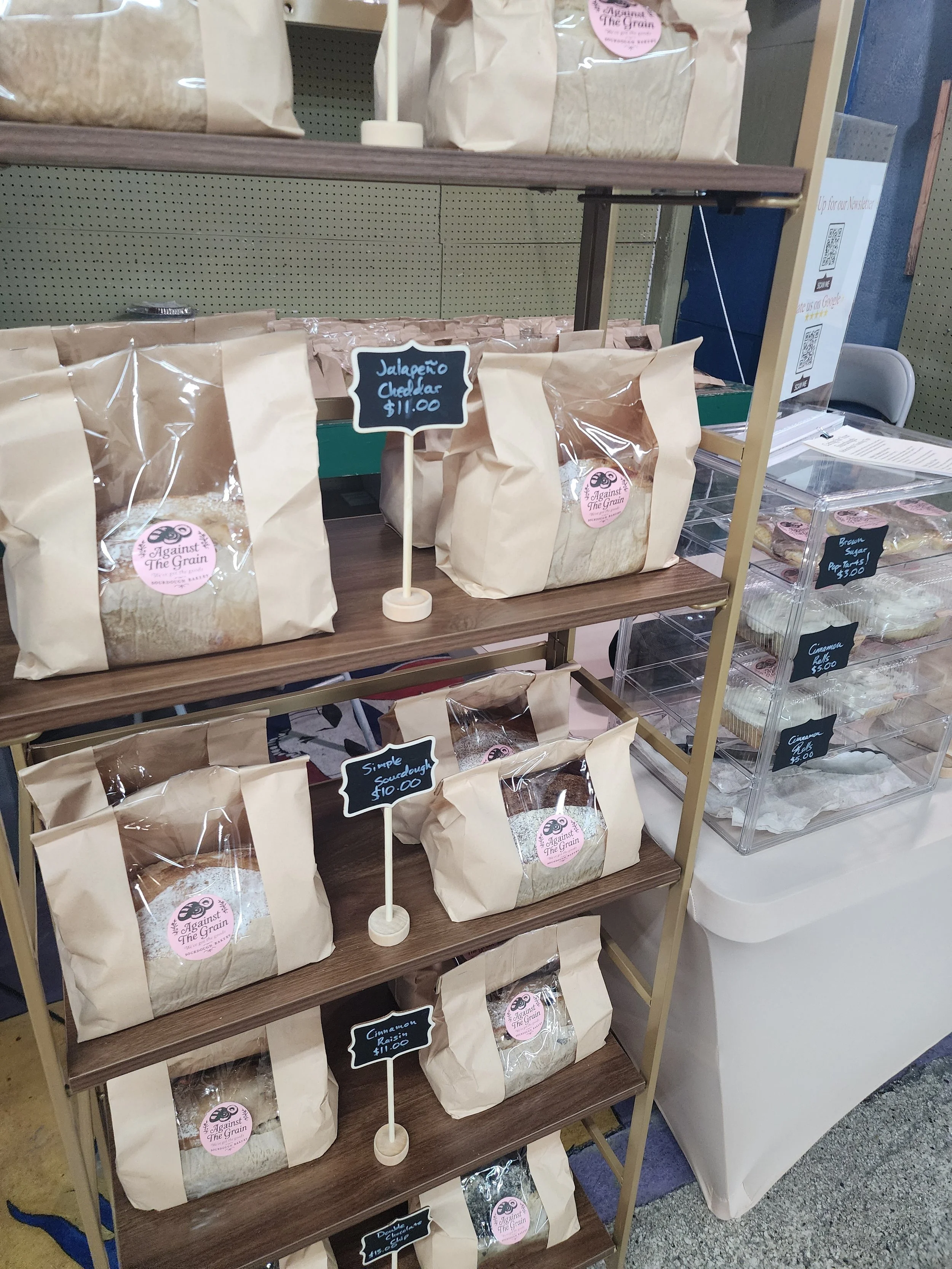 Pantry shelf containing beige paper bags of different types of baked goods with small chalkboard signs labeling the items, including jalapeño cheddar bread priced at $11 and cinnamon raisin bread priced at $11, with a packaged snack on the right side