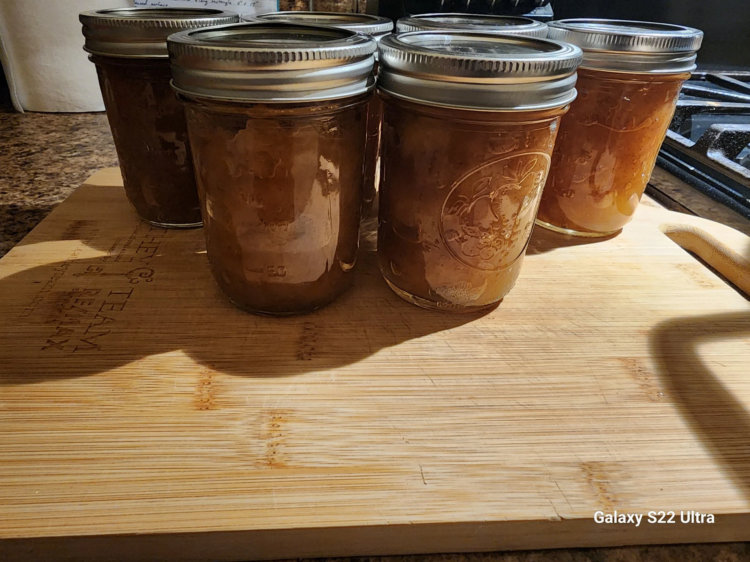 Multiple jars with silver lids are placed on a wooden surface. The jars appear to contain a brown substance, possibly homemade preserves or sauce. There is a stove and a piece of paper or cloth in the background.
