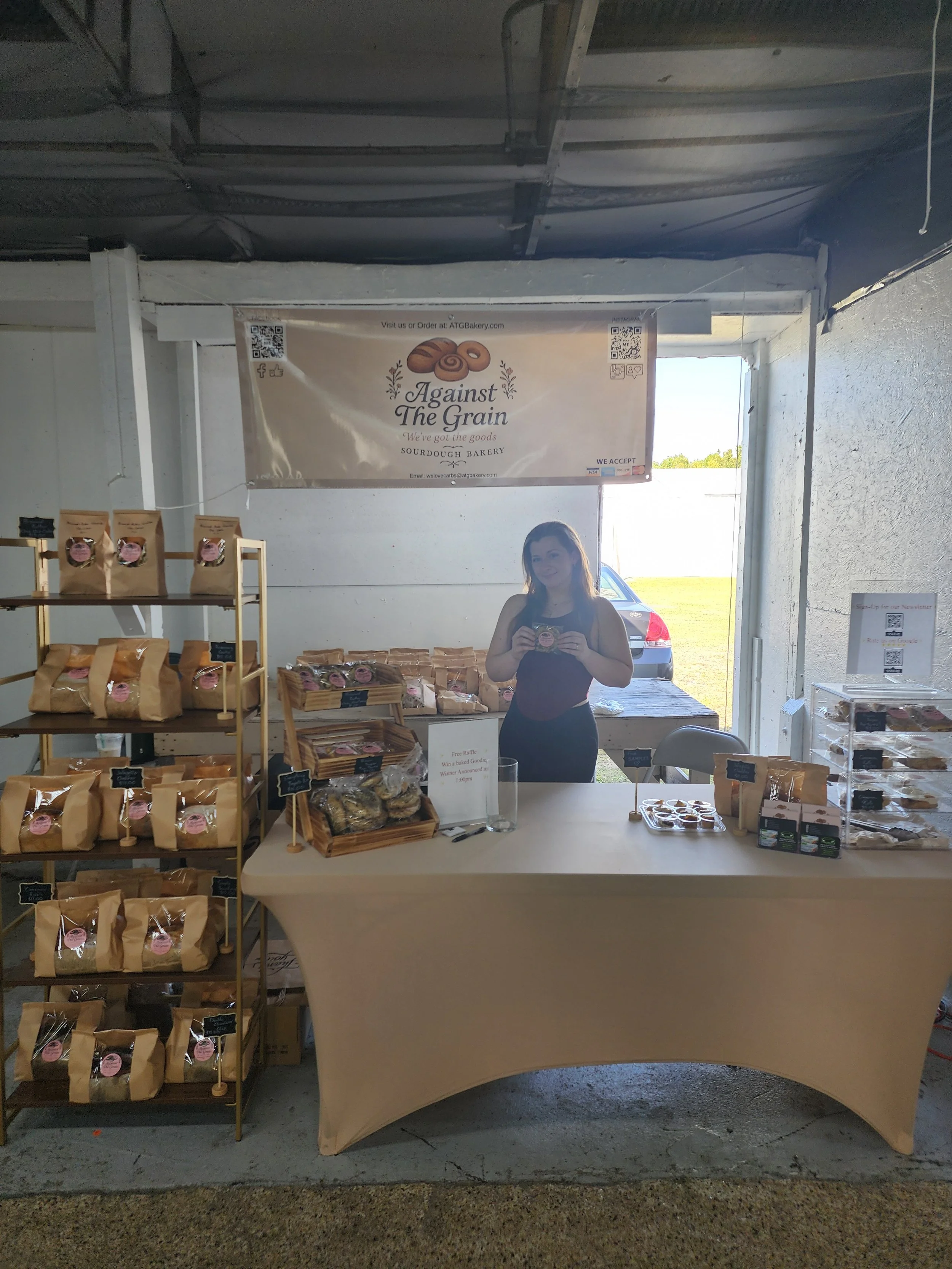 A woman at a bakery booth holding a package of baked goods, with shelves of packaged bread and baked items for sale, a banner for 'Against The Grain Sourdough Bakery,' and a bright outdoor background.