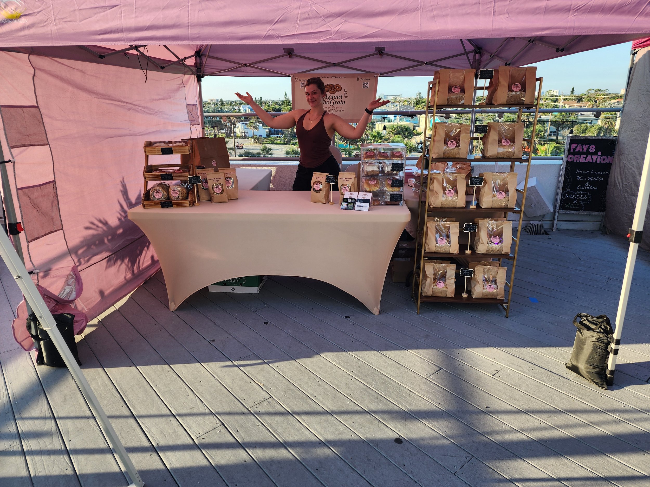 A woman standing behind a booth at an outdoor market, with shelves of baked goods and a pink canopy overhead, on a rooftop with a city view in the background.