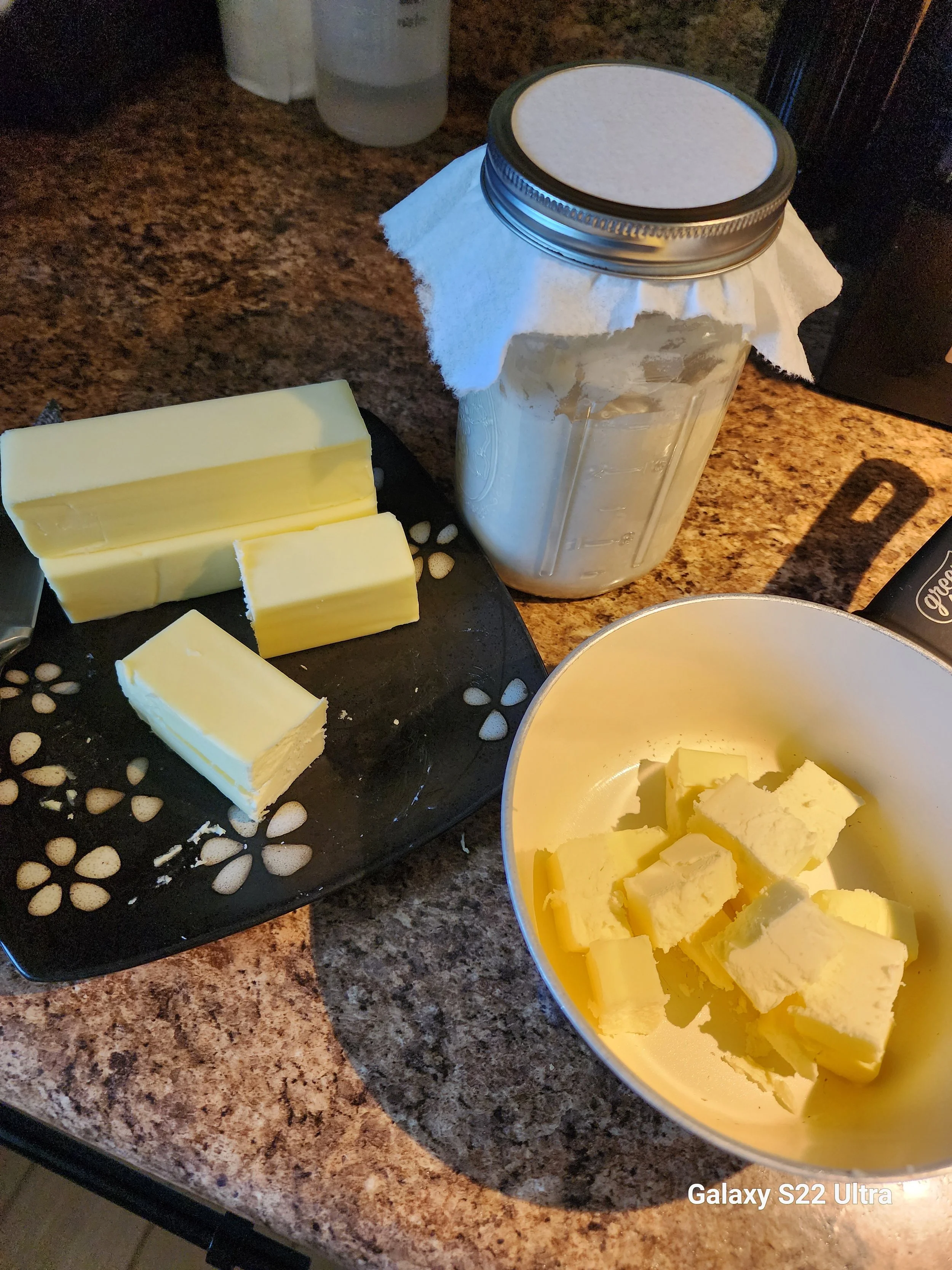 Blocks of butter on a black plate and in a yellow bowl on a kitchen counter, with a jar and bottles in the background.