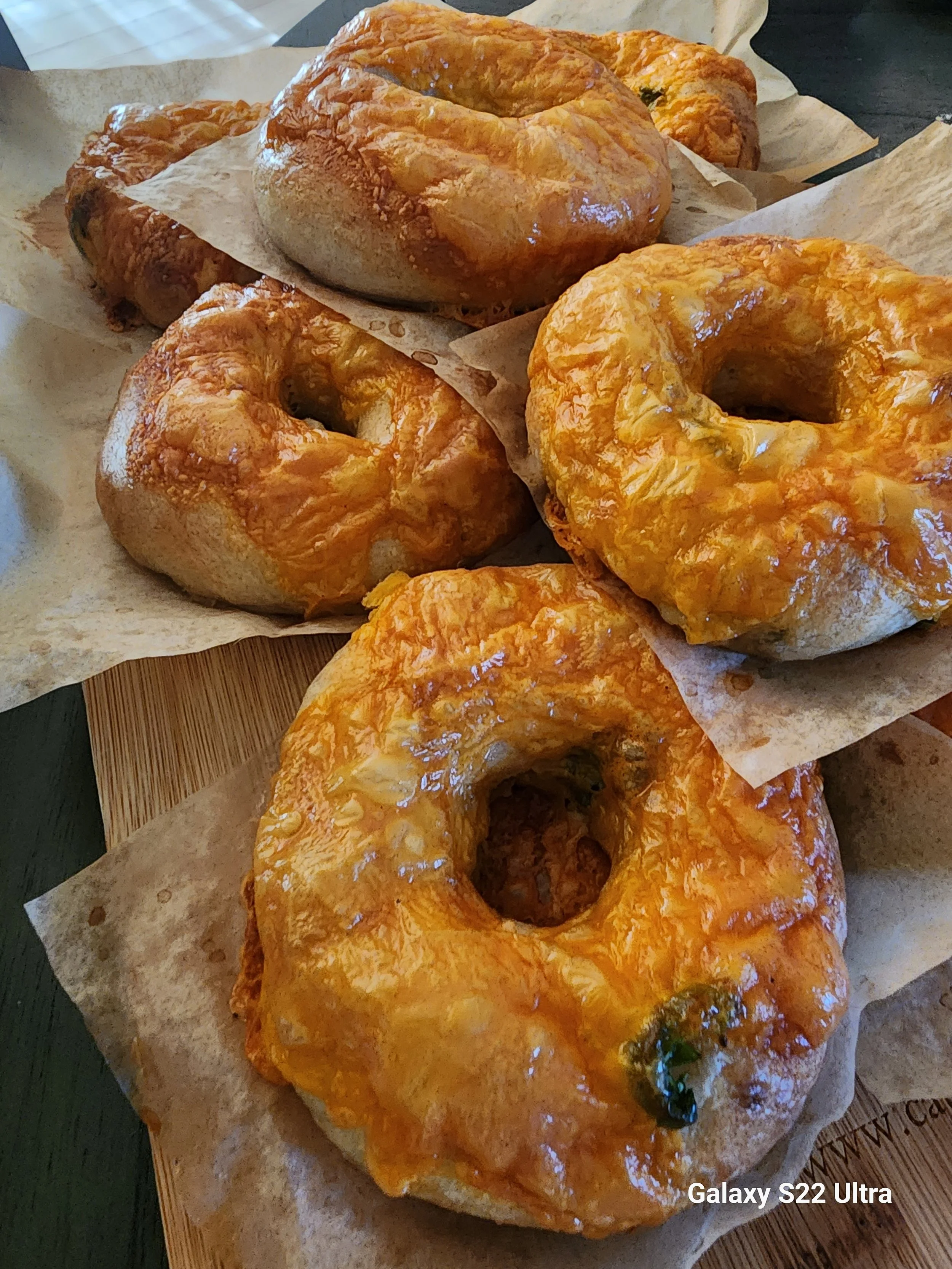 Close-up of three cheese bagels on parchment paper, different shades of melted cheese on top, placed on a wooden surface.