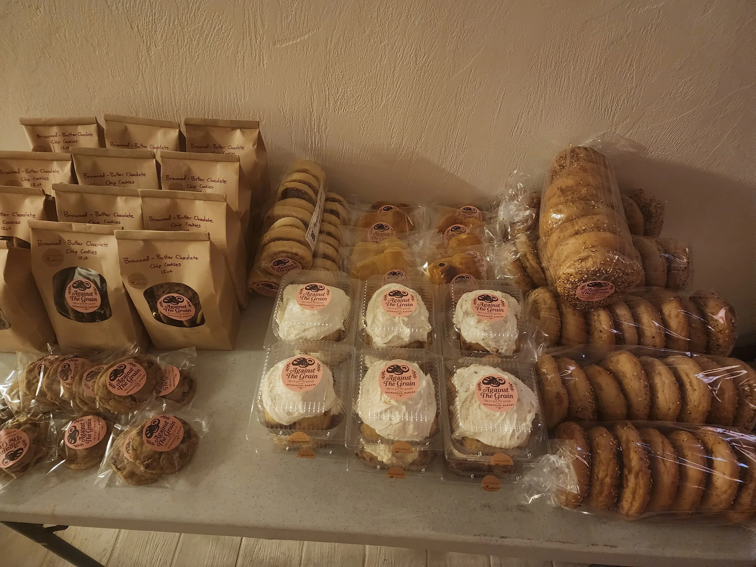 Shelf with packaged baked goods including cookies, cookies in bags labeled 'Browned-Butter Chocolate Chip Cookies,' and slices of bread. The items are wrapped in plastic and displayed on a white surface.