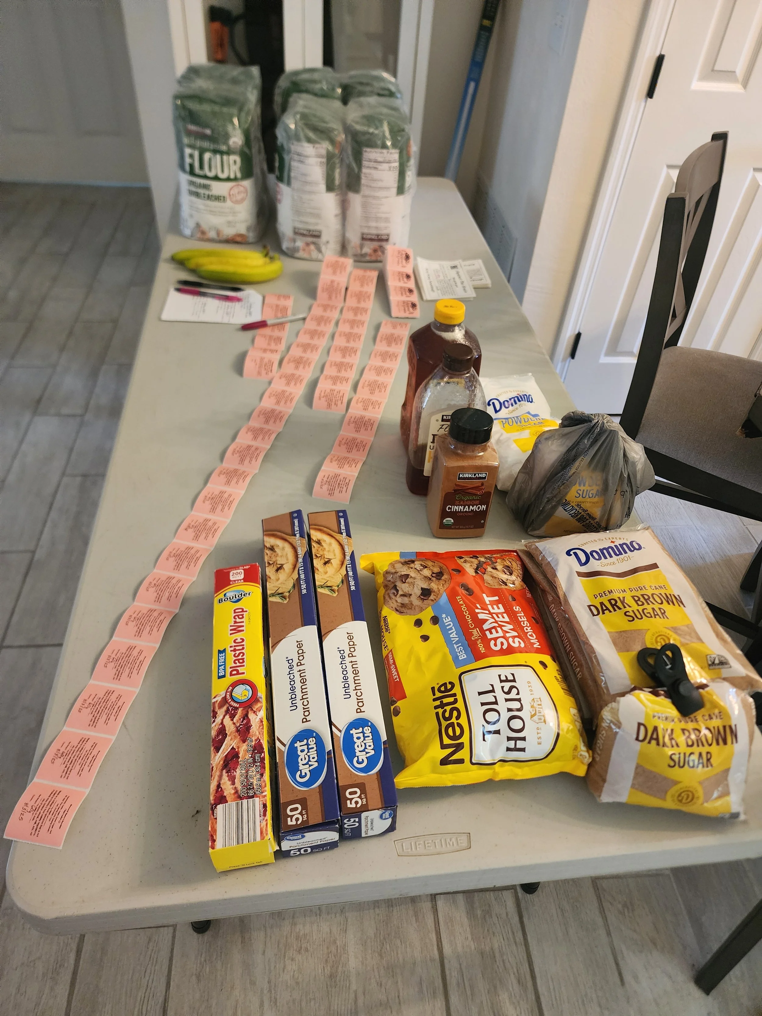 A table with various grocery items including flour, bananas, baking ingredients, and packaged cookies or snacks, with coupons or receipts laid out alongside.