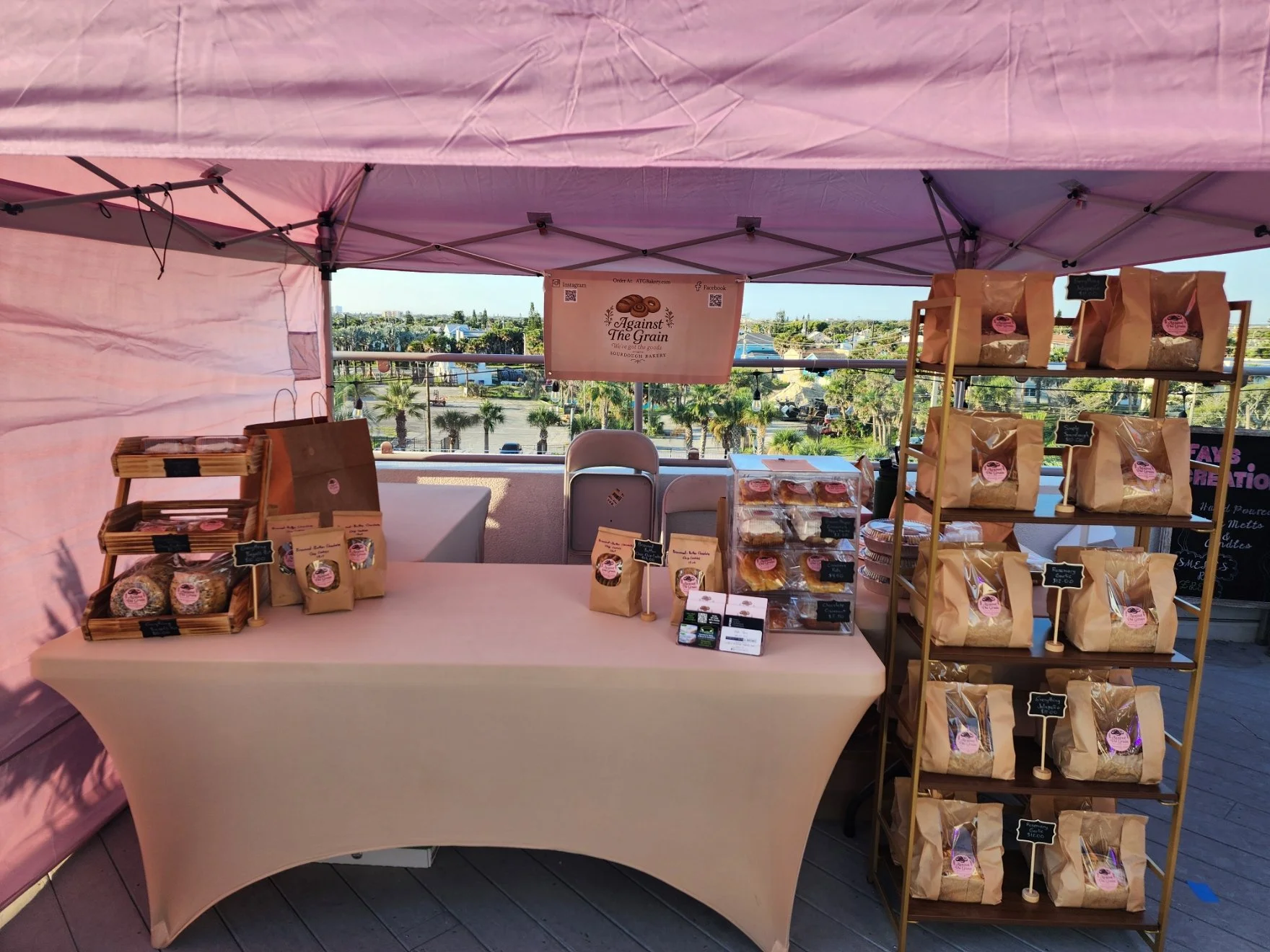 A bakery booth with baked goods and bread in paper bags on display shelves and a table, with a pink canopy, set outdoors with trees and a parking lot in the background.