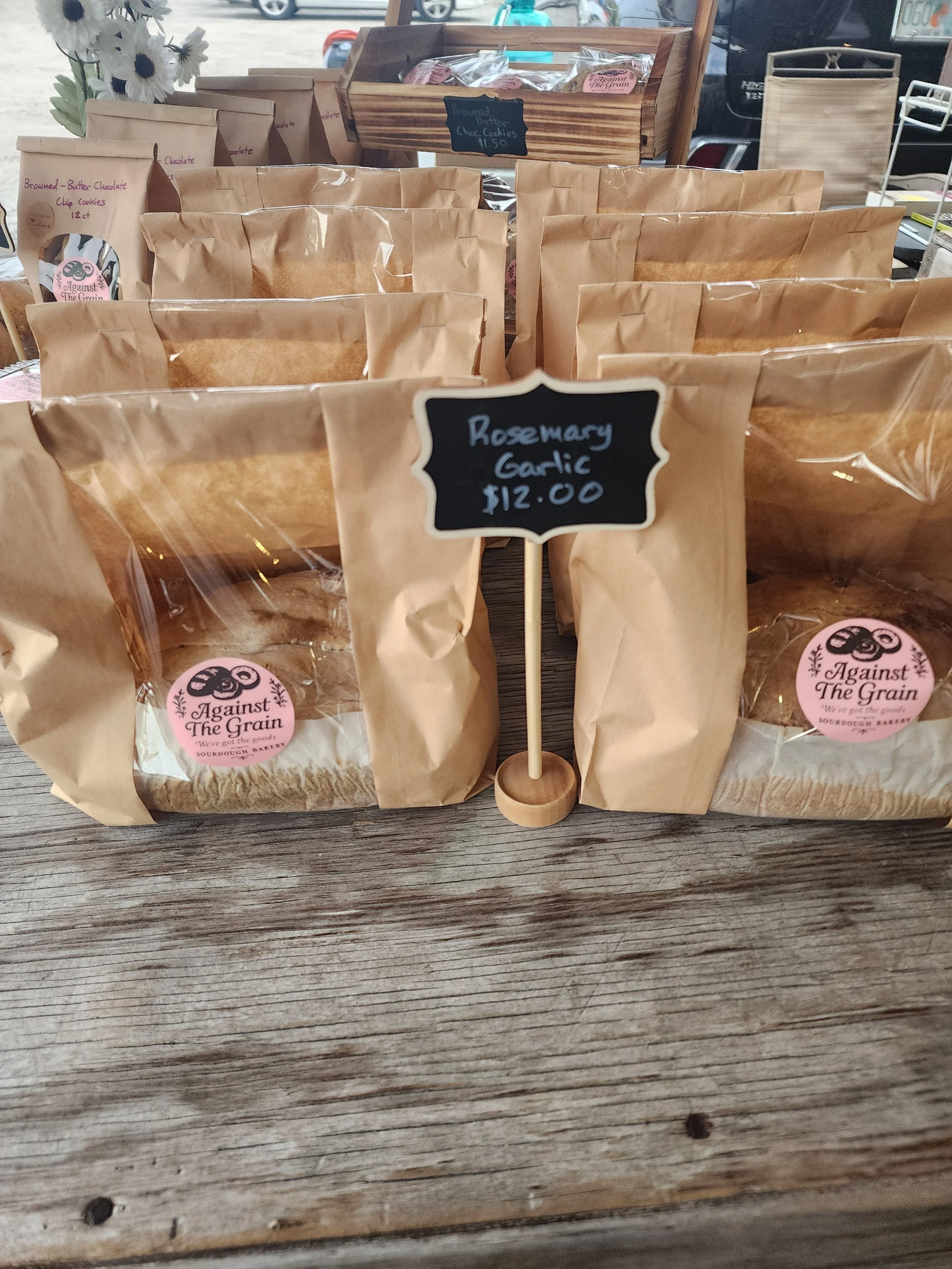 Display of brown paper bags with a sign that reads 'Rosemary Garlic $12.00' at an outdoor market or shop table.
