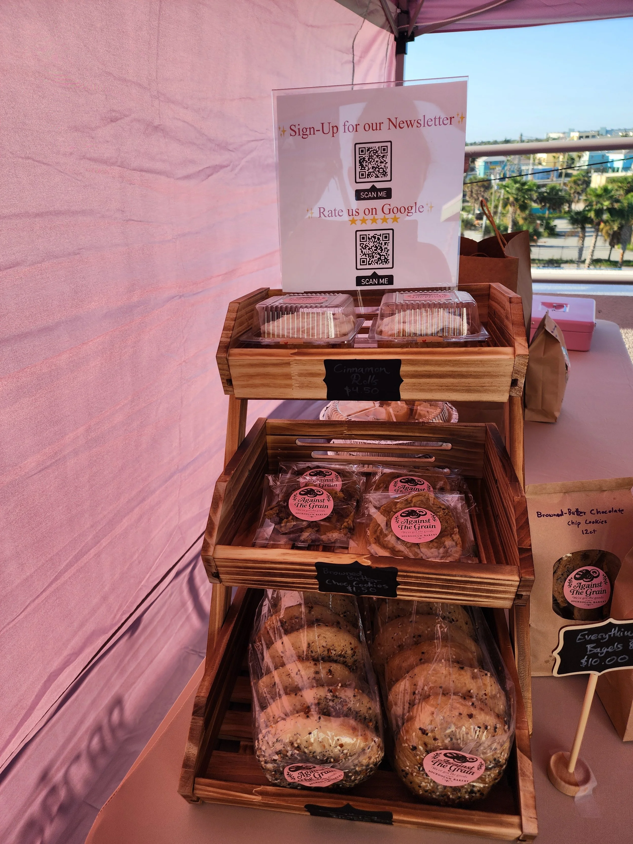 A display stand with baked goods including cookies and rolls, labeled with signs. Sign-up sheet and QR codes are placed on top of the stand for newsletter subscription and Google reviews.