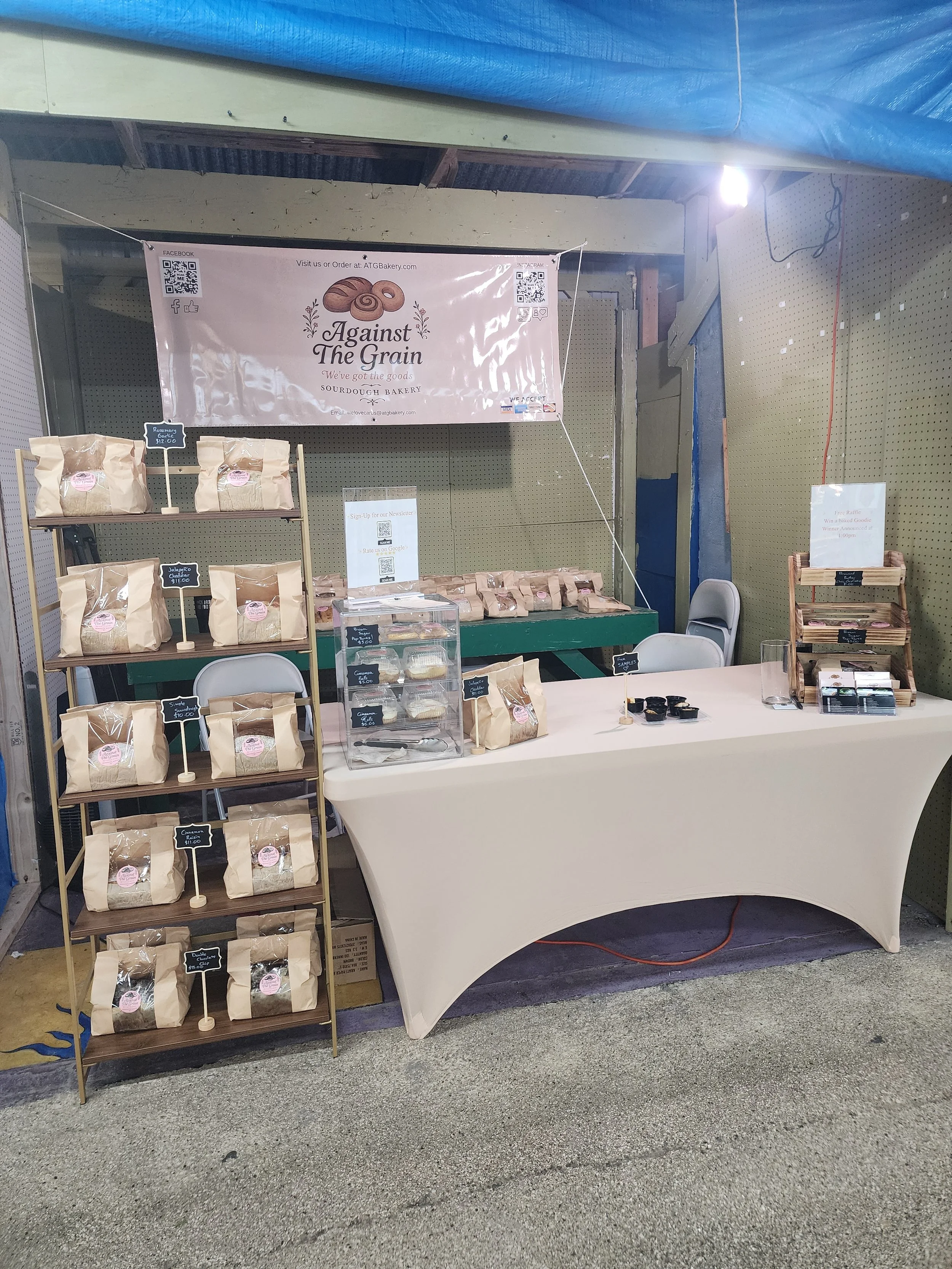A bakery booth at a fair, featuring bags of bread, cupcakes, and baked goods with signs and a large banner reading 'Against the Grain'.