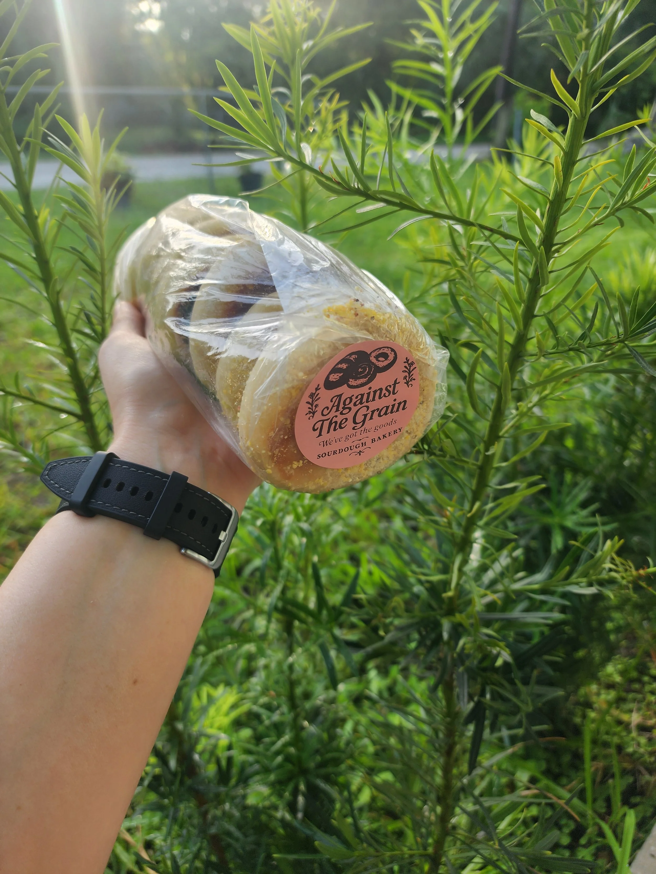 A person holding a packaged baked good with a label that reads "Against The Grain" in front of a green leafy plant. The person is wearing a black watch.