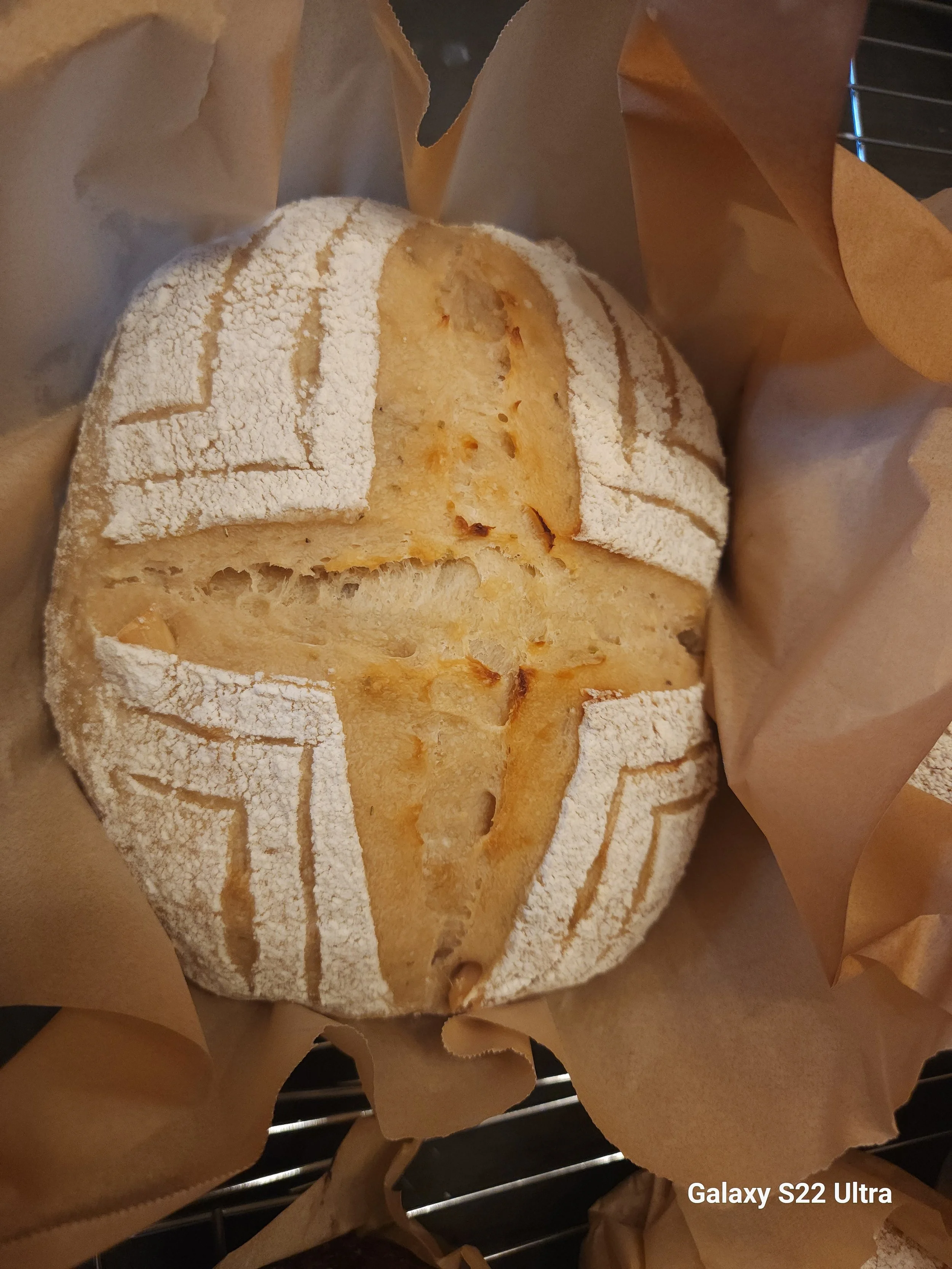 A round loaf of bread dusted with flour, wrapped partially in brown paper. The loaf has a golden crust with a scoring pattern on top.
