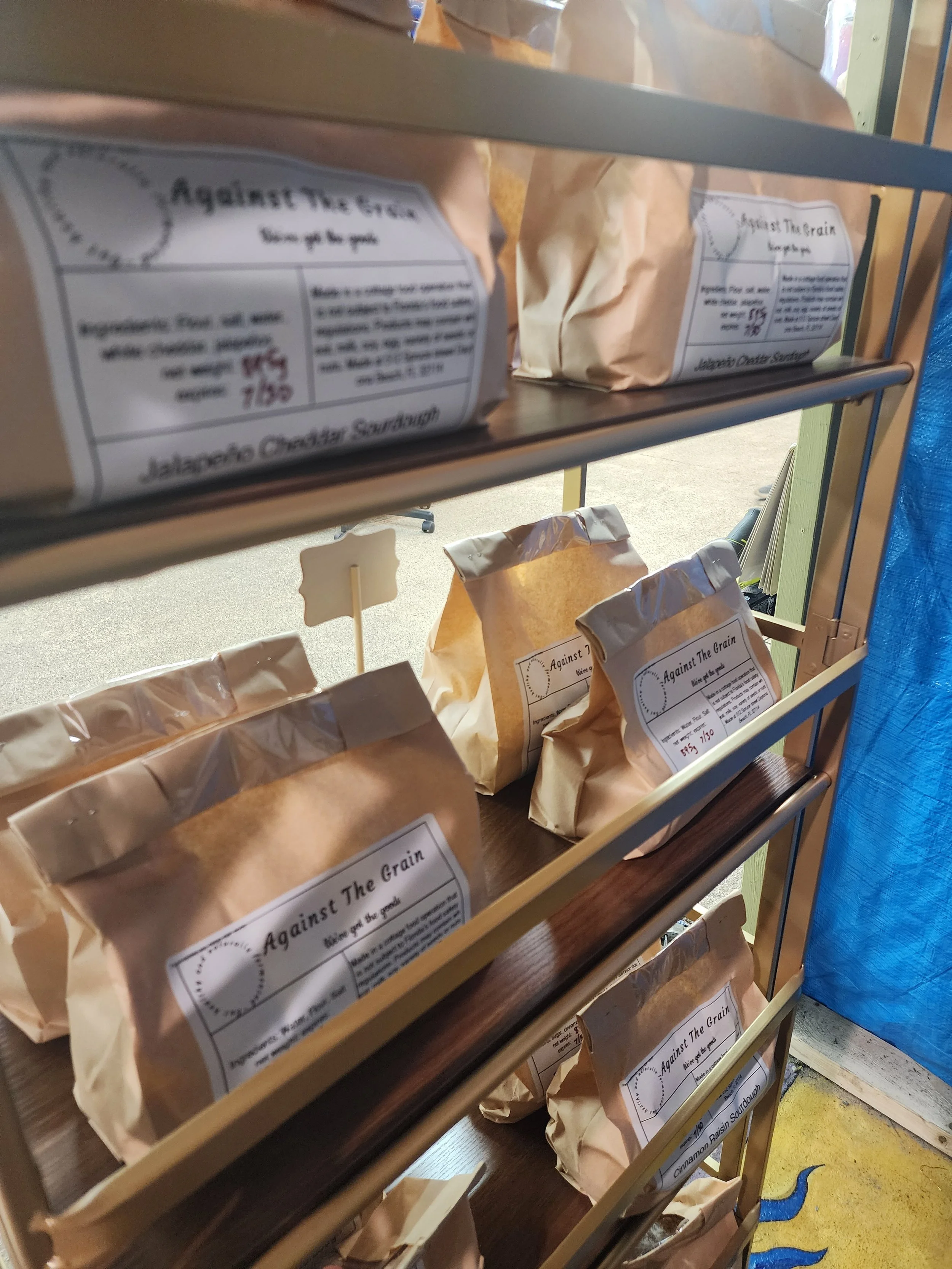 Shelf with bags of baking ingredients in paper packaging labeled "Against The Grain" at a store or bakery.