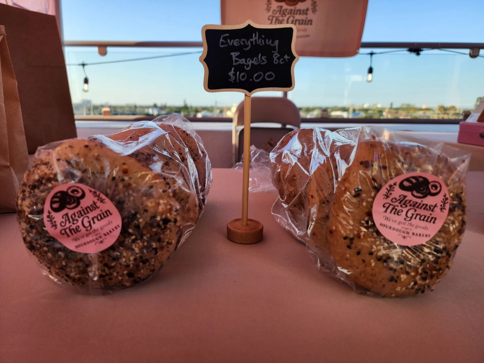 Two bags of seed-covered bagels in clear plastic packaging on a pink table. A small sign says, 'Everything Bagels $10.00.' In the background, there is a window and a sign for 'Against The Grain' bakery.