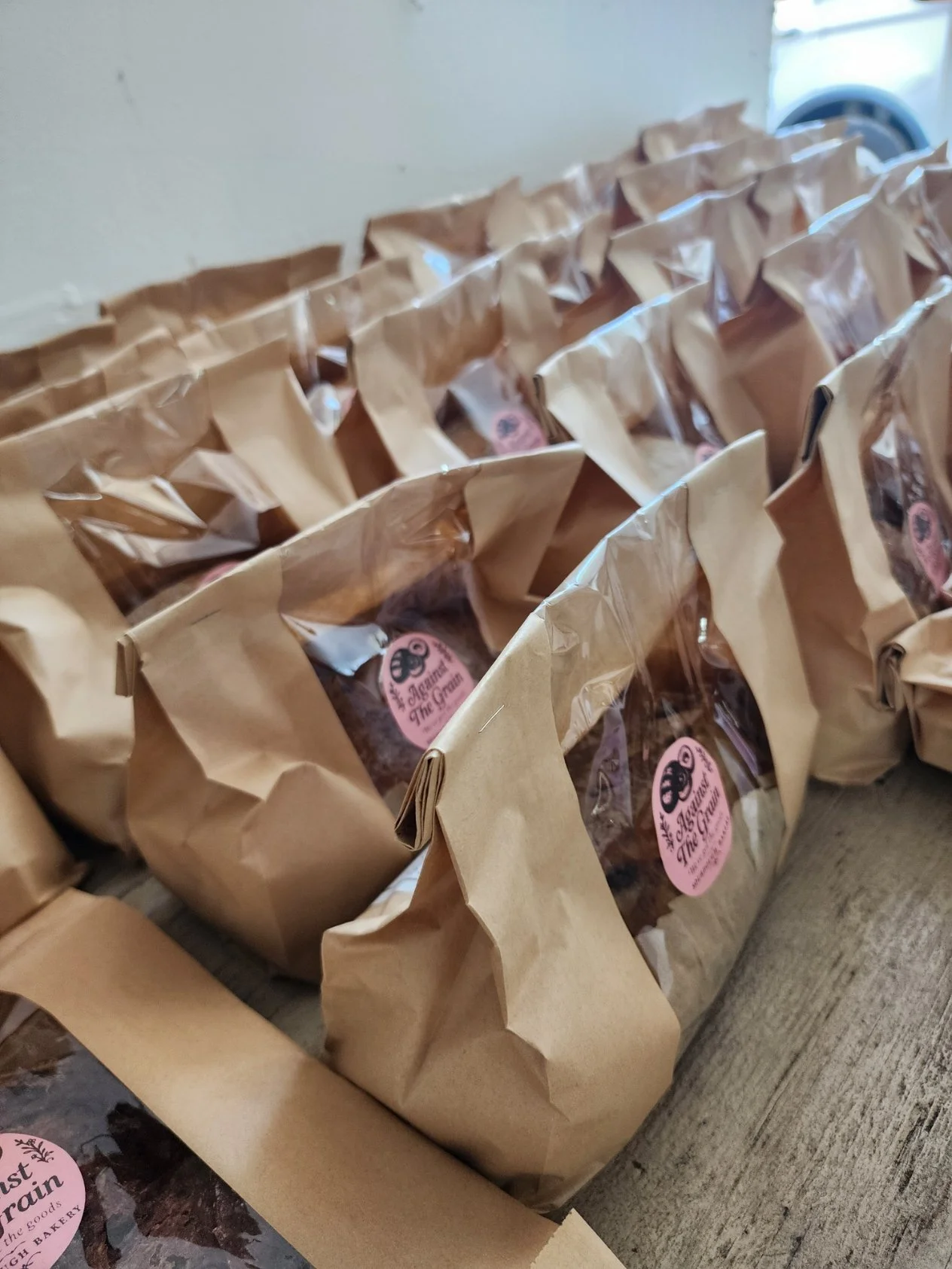 Multiple brown paper bags with transparent windows and pink stickers, arranged on a table near a white wall and a washing machine.