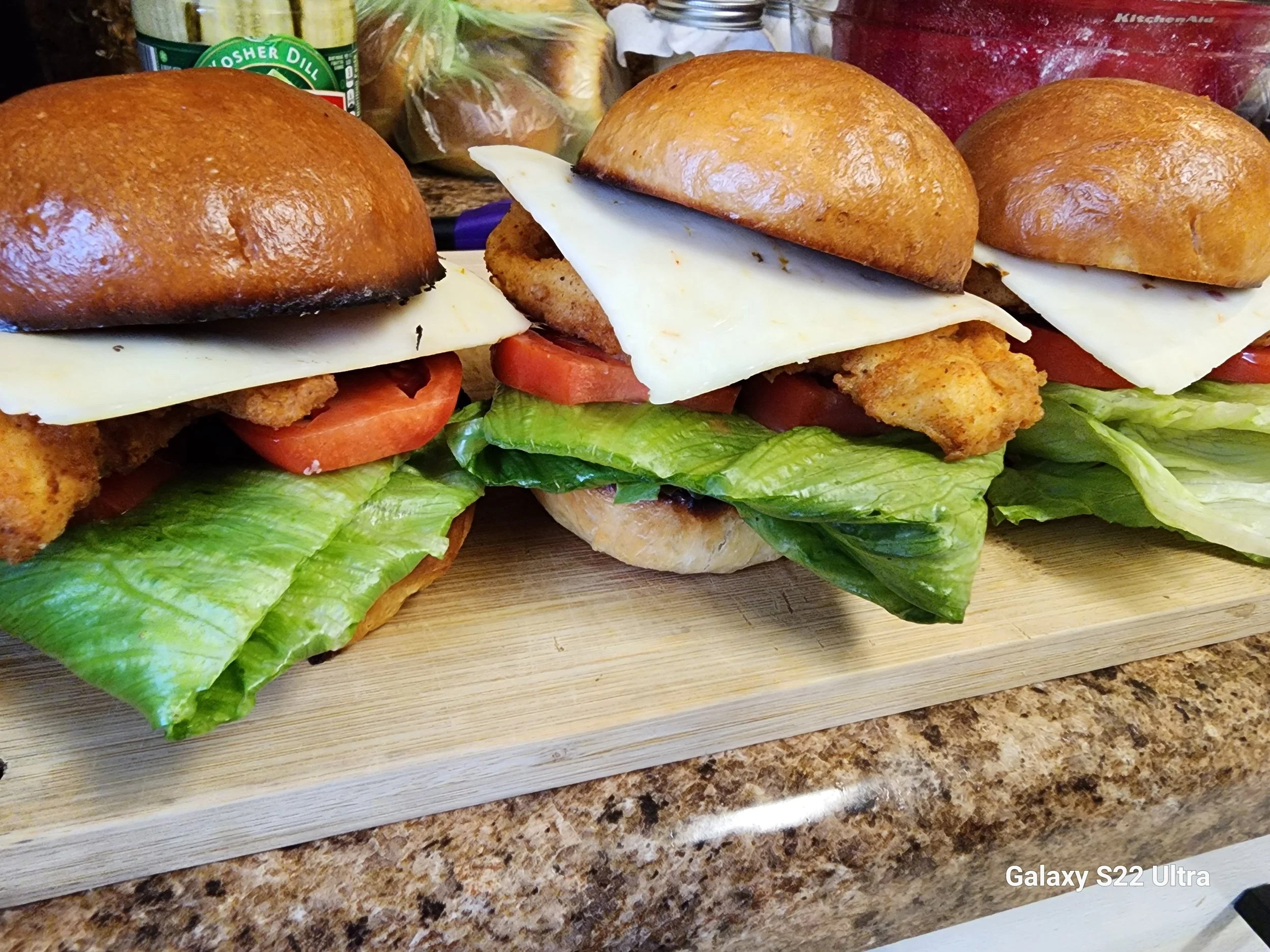 Three fried chicken sandwiches on toasted buns with lettuce, tomato, cheese, and fried chicken fillets, placed on a wooden cutting board on a kitchen countertop with various food items in the background.