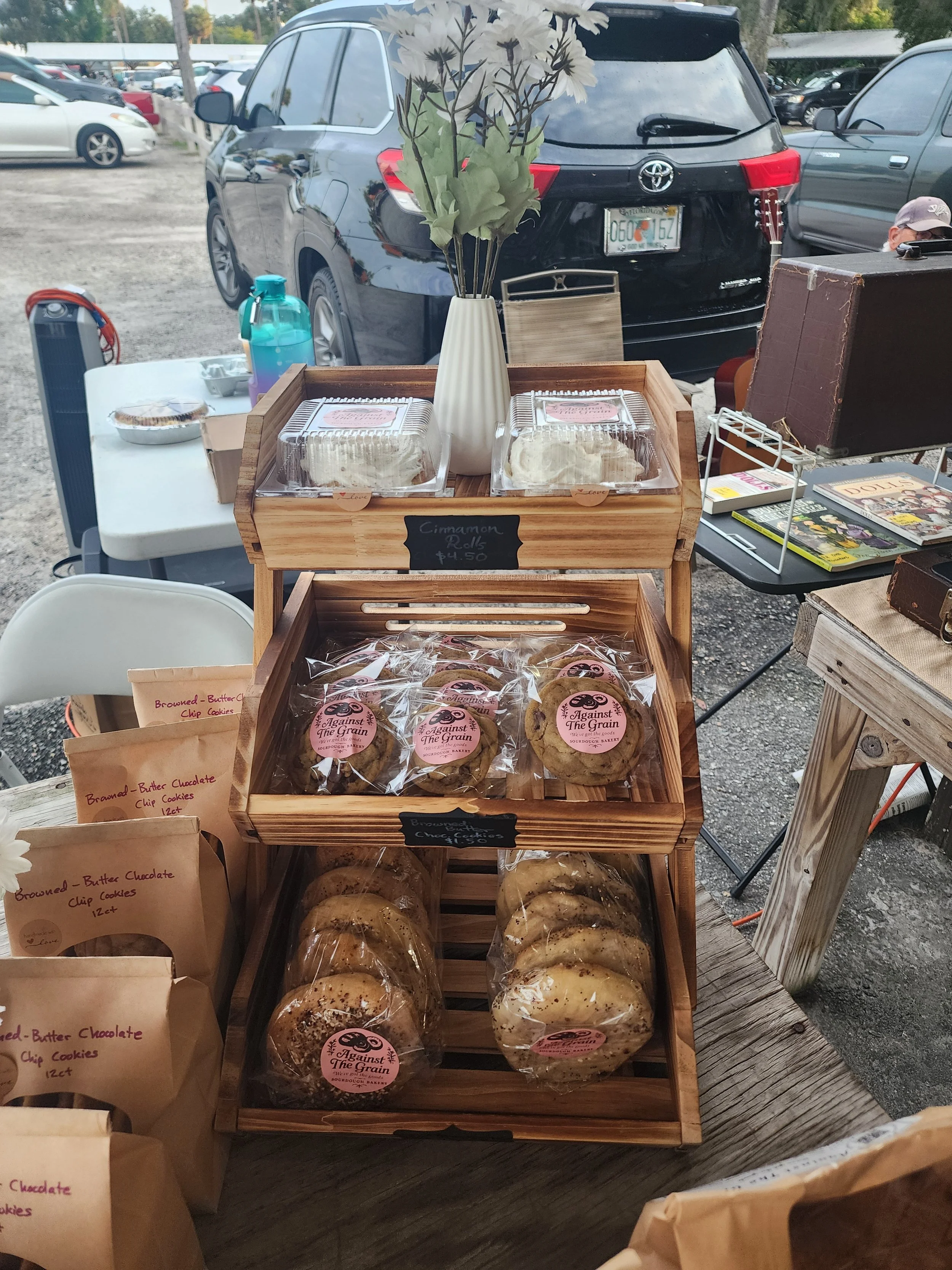 Display of baked goods at an outdoor market, including cookies labeled "Against The Grain," with a vase of artificial flowers in the background and a variety of other items on the table.