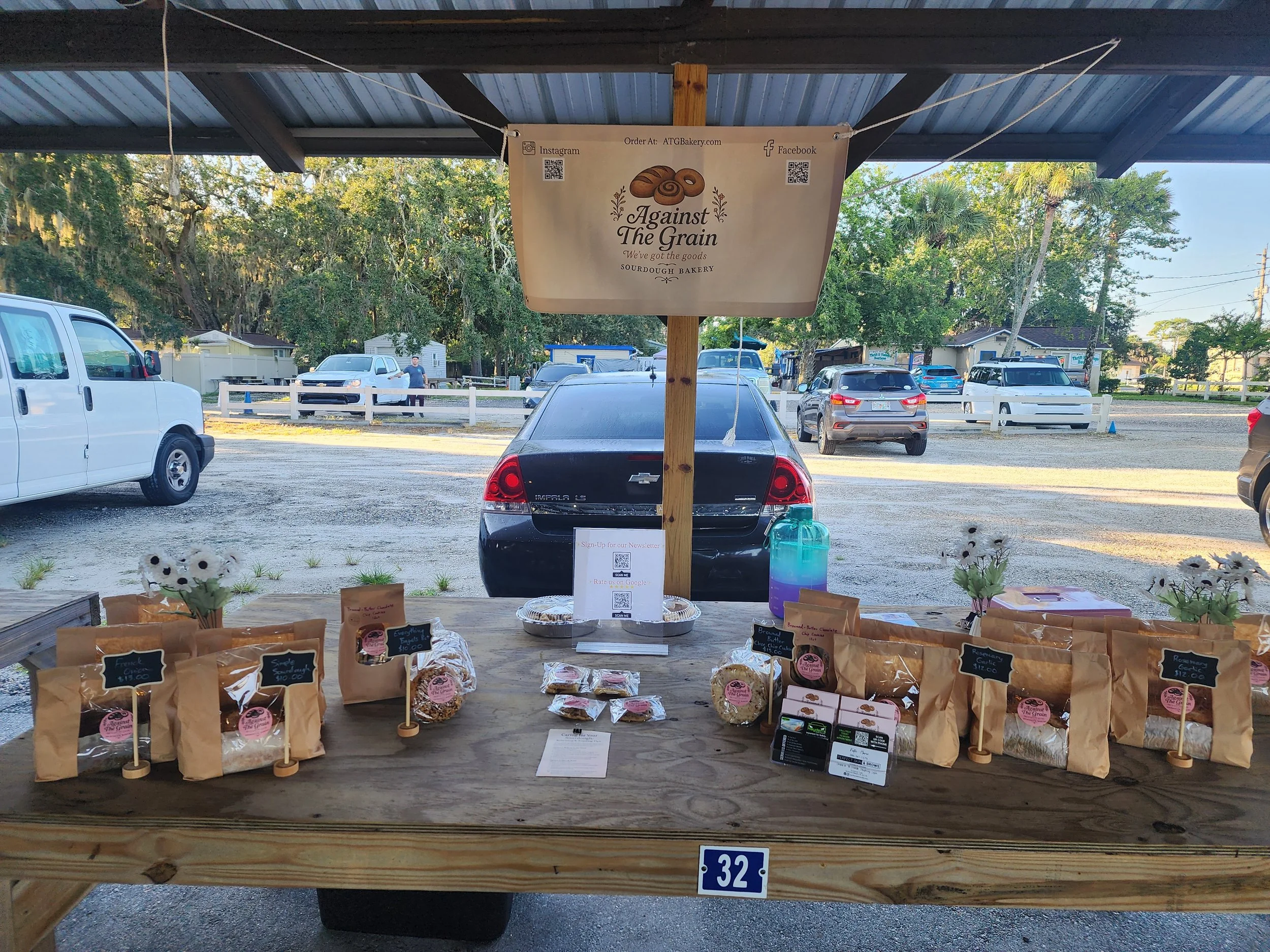 A market stall with baked goods in paper bags on a wooden table, with a sign for Against The Grain Sourdough Bakery and cars parked in the background.