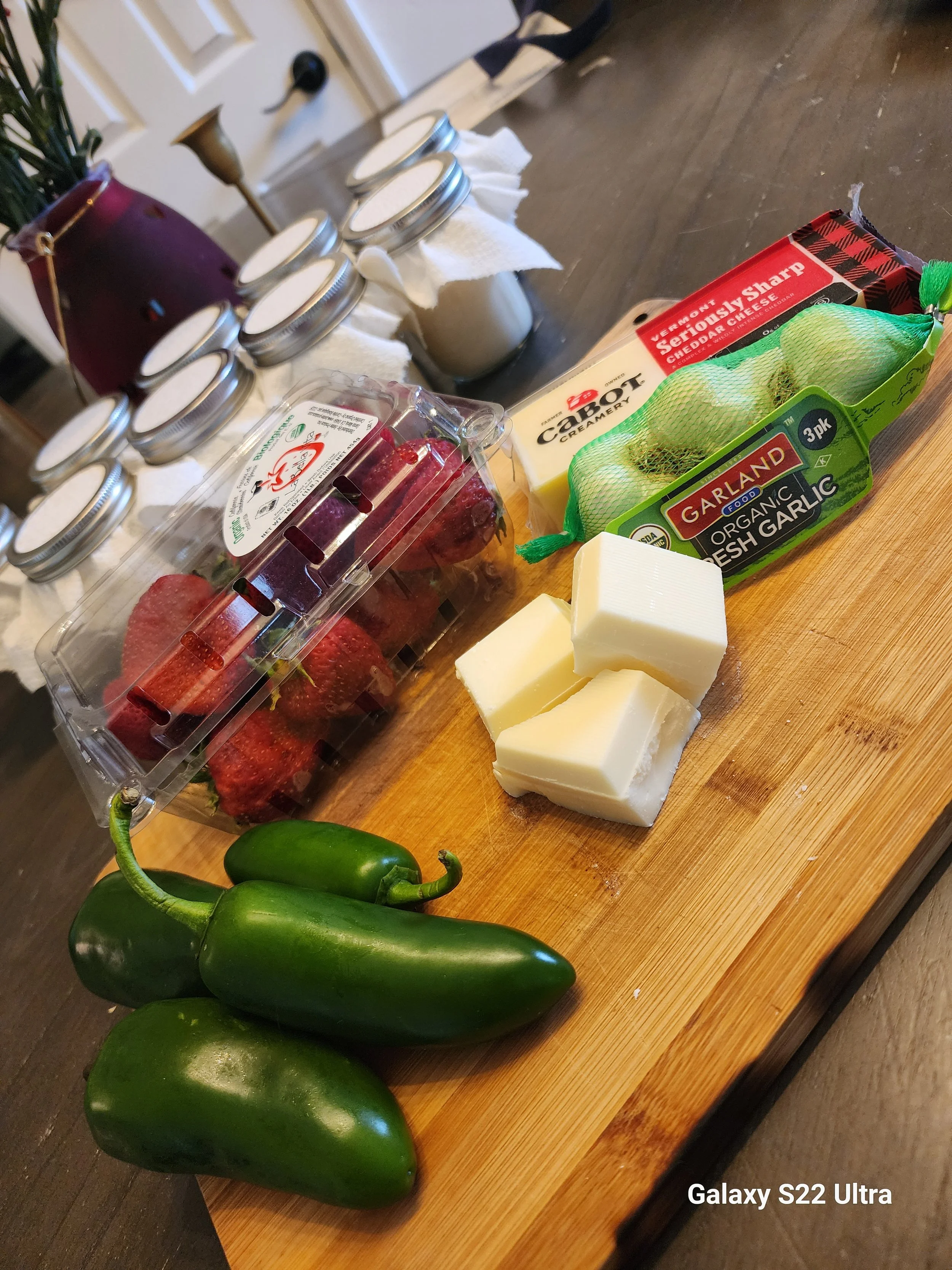 Green jalapeño peppers, a block of cheese, strawberries in a plastic container, a bag of organic garlic, a box of sharp cheddar cheese, and jars on a wooden table in a kitchen.