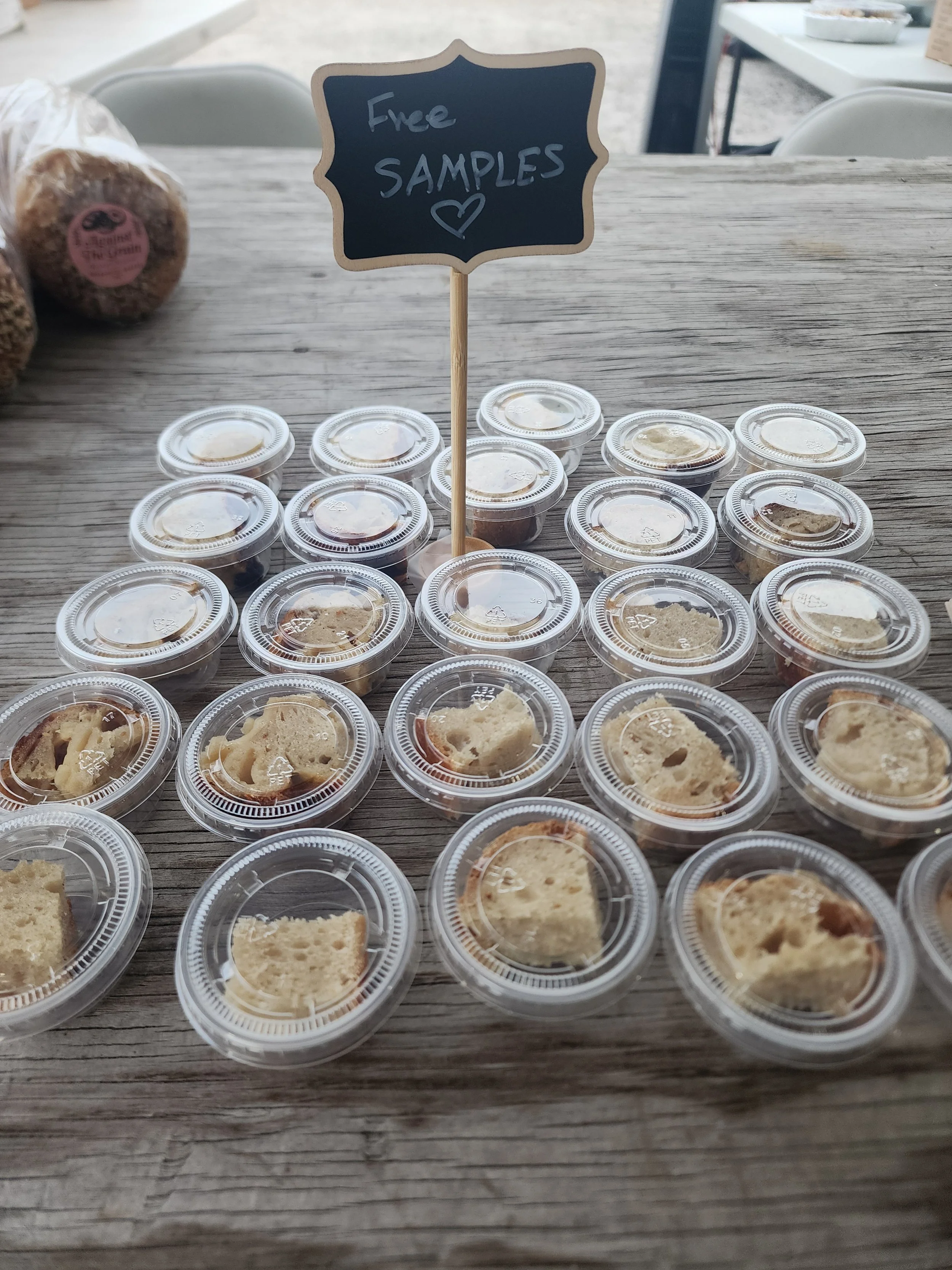 Small clear plastic containers of bread samples arranged on a wooden table with a sign that says 'Free Samples' and a heart drawing.