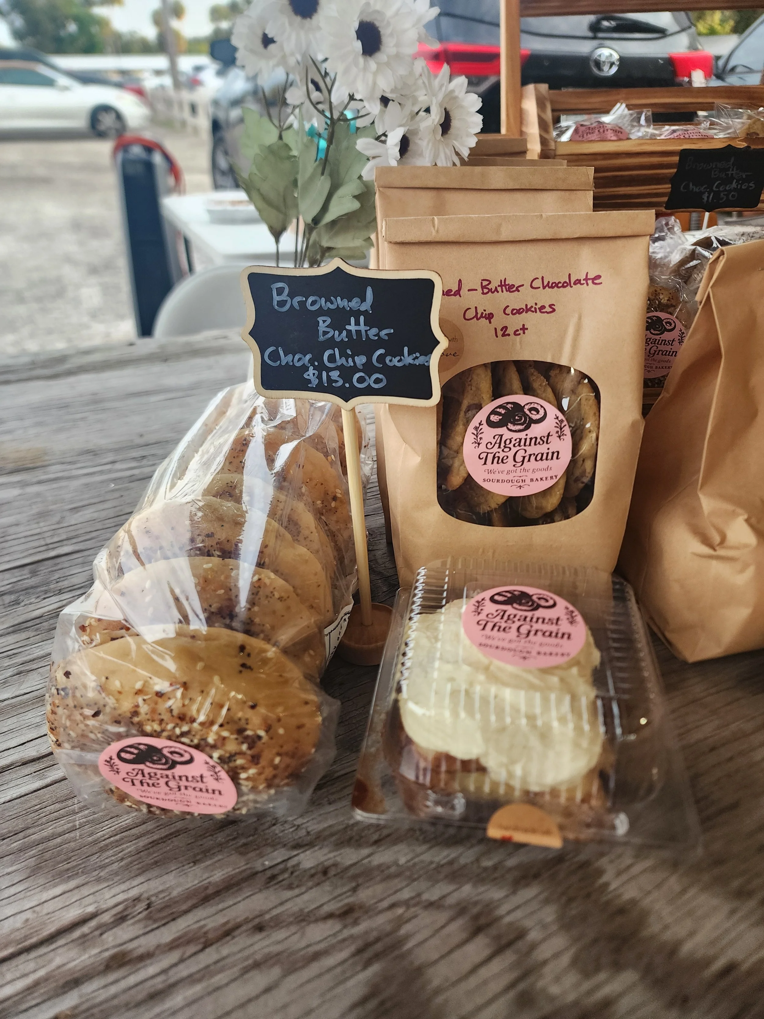 Baked goods, including cookies in clear plastic packaging and a cake, displayed on a wooden table with a small bouquet of white flowers, at an outdoor market