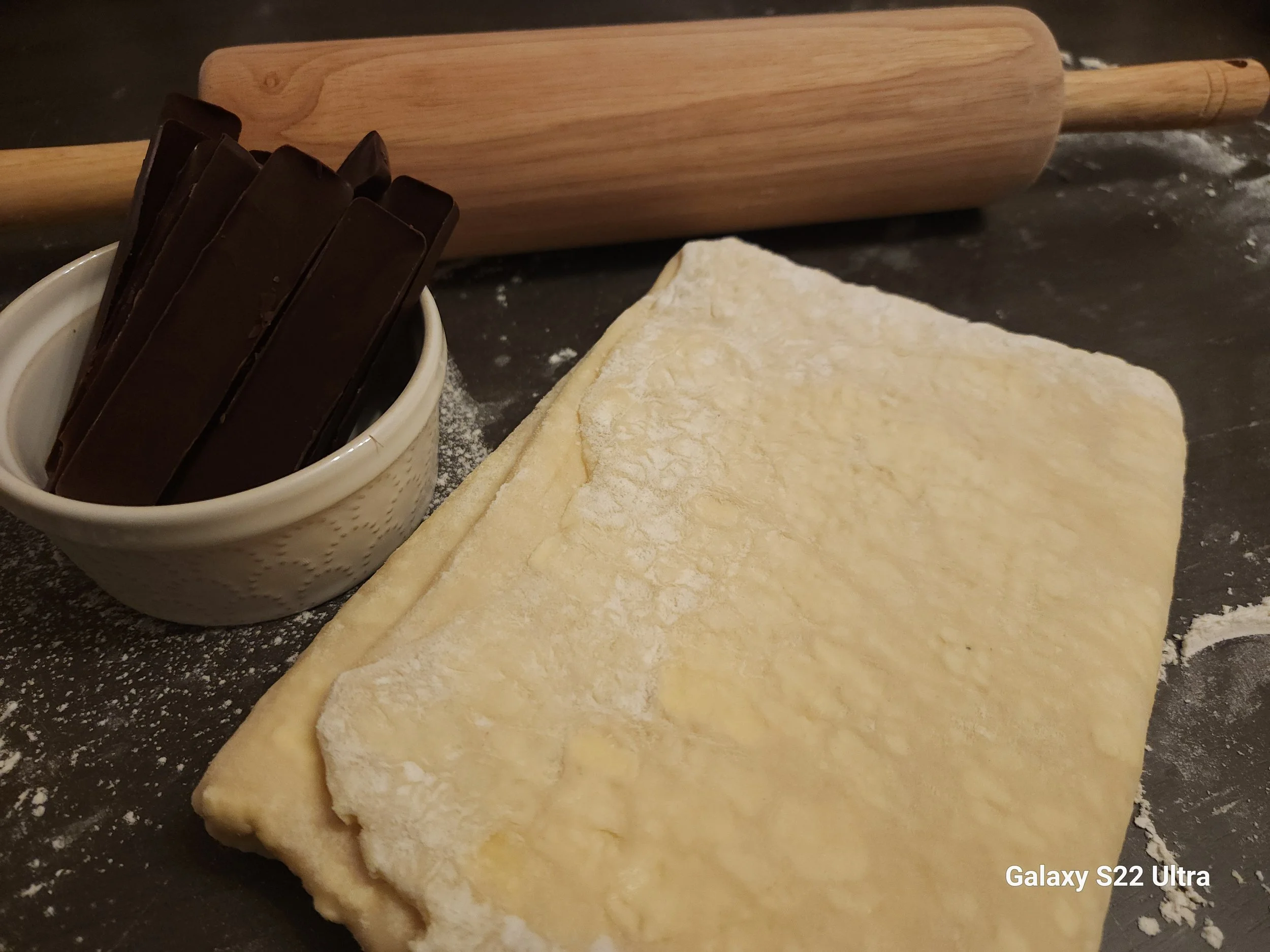 A rolling pin, a bowl of chocolate, and a partially rolled out piece of dough on a black countertop with flour.