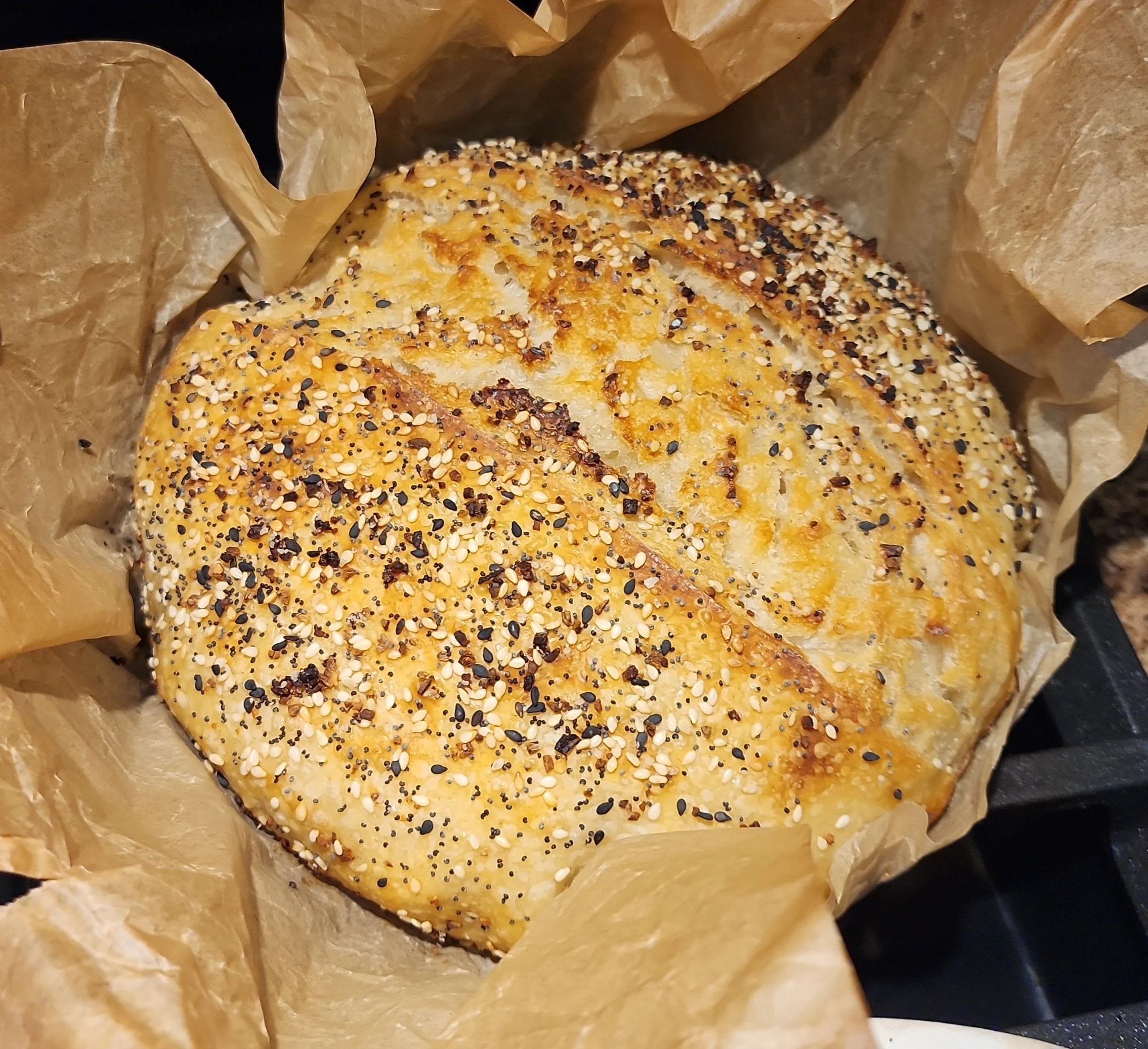A loaf of bread topped with black and white sesame seeds, partially wrapped in brown parchment paper.