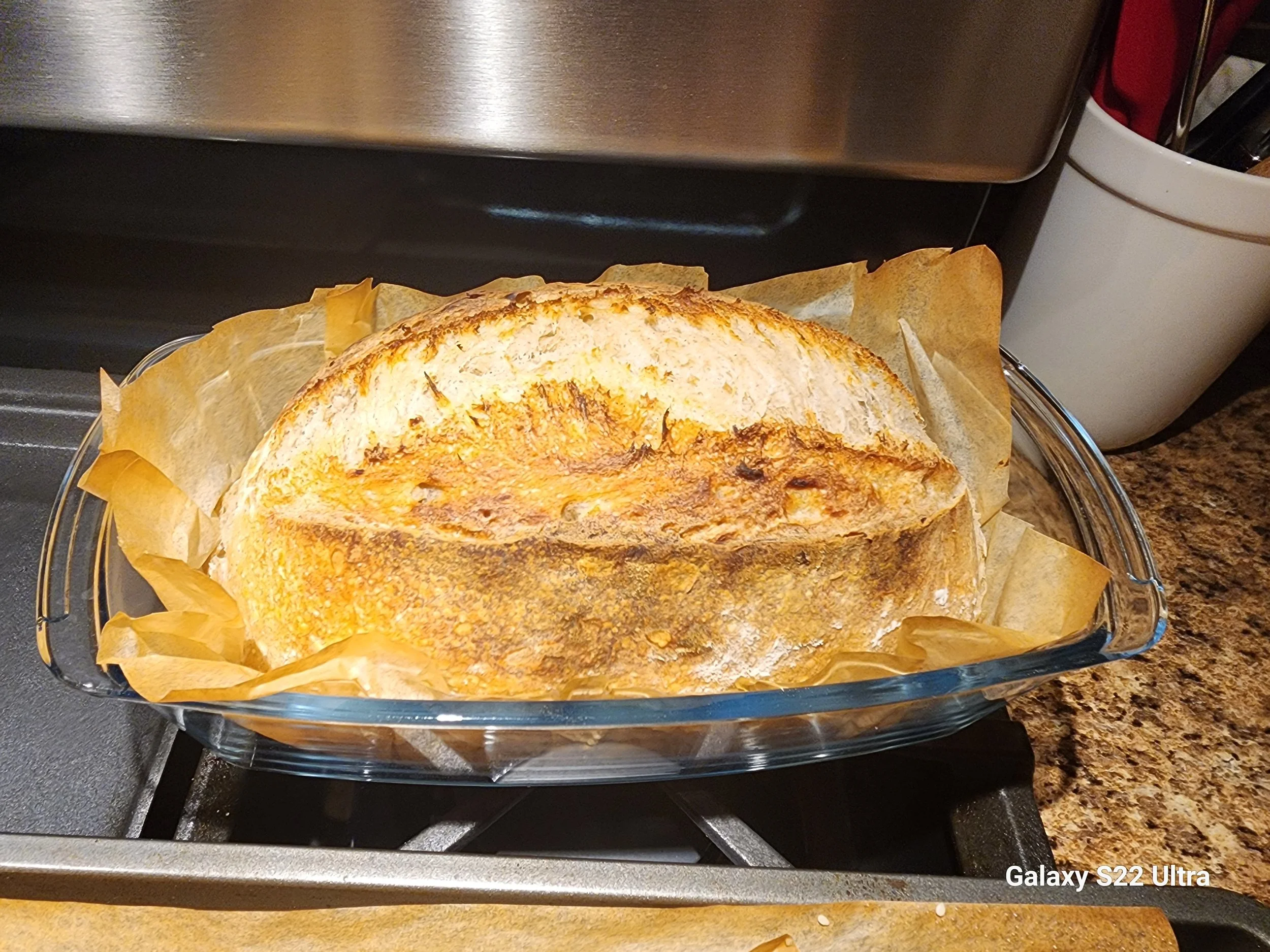 Baked loaf of bread in a glass baking dish lined with parchment paper, placed on a stovetop. The bread has a golden-brown crust.