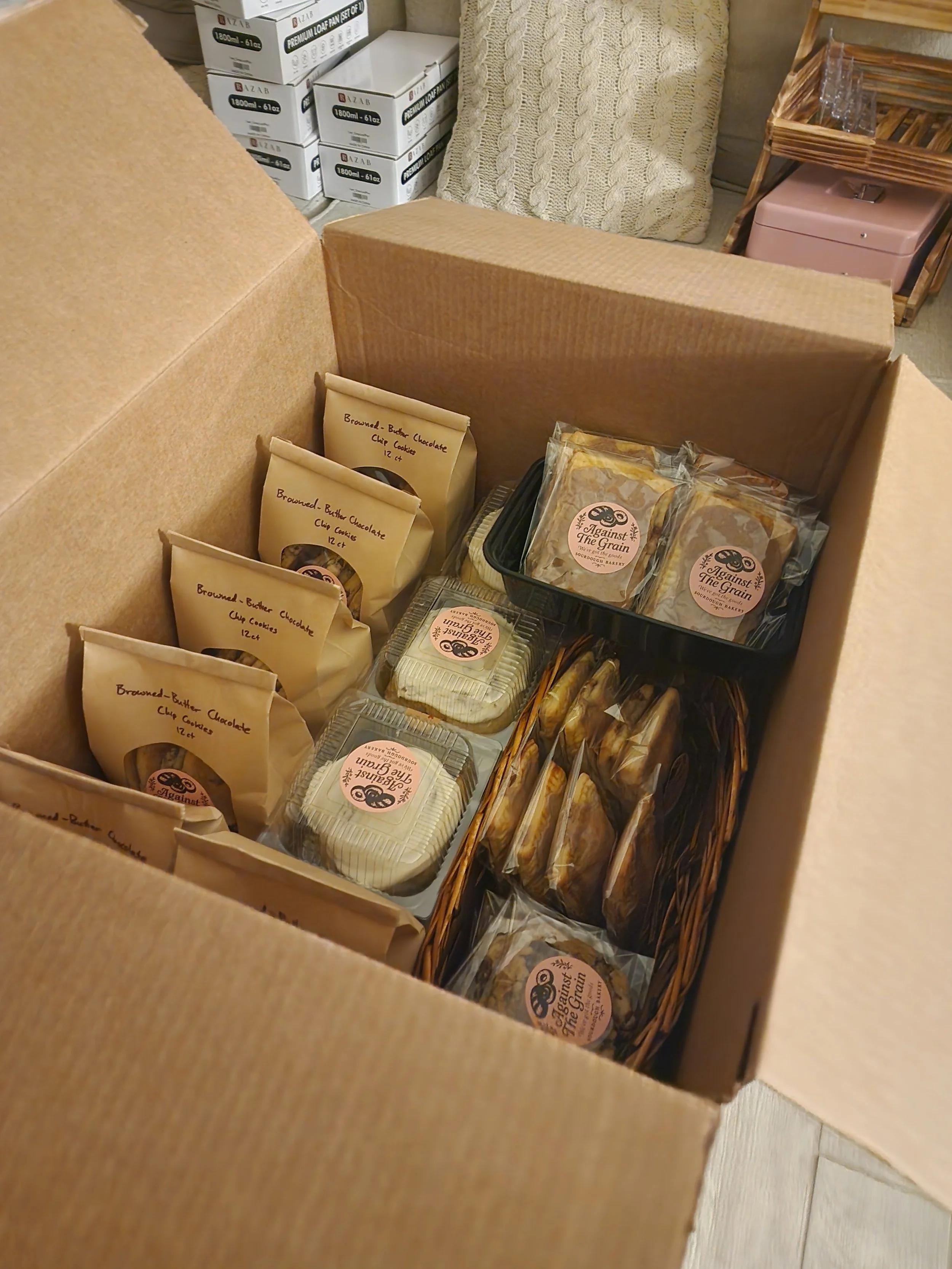 A cardboard box containing packages of baked goods, including cookies labeled 'Browned Butter Chocolate Chip Cookies,' with a background of boxes and a cushioned pillow.