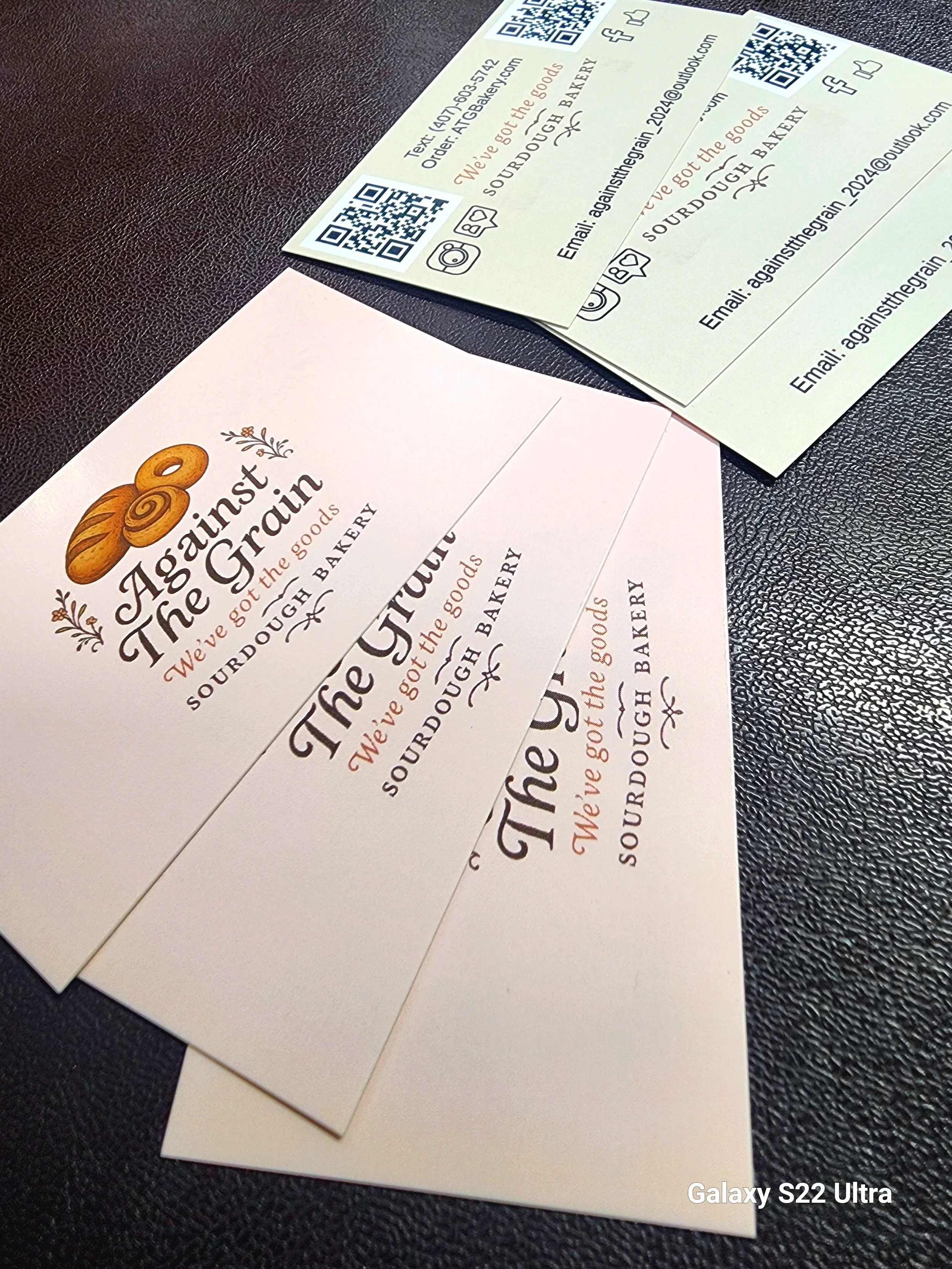 Close-up of business cards for Against the Grain bakery, featuring a logo with a croissant illustration and text that says "Against the Grain, We've got the goods". The cards are on a textured black surface.